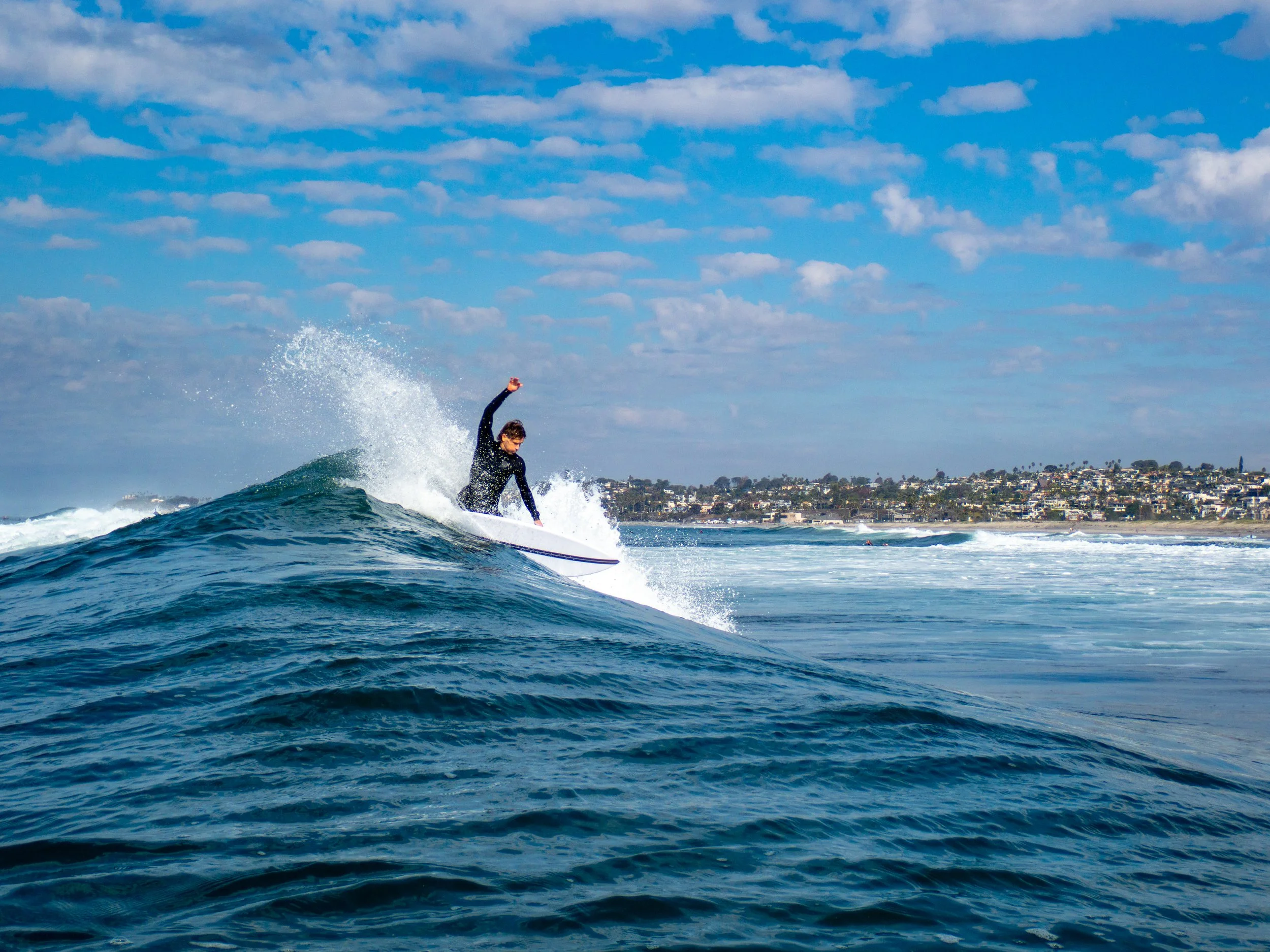 A person riding a wave on a surfboard in the ocean with a coastal town in the background under a partly cloudy sky.