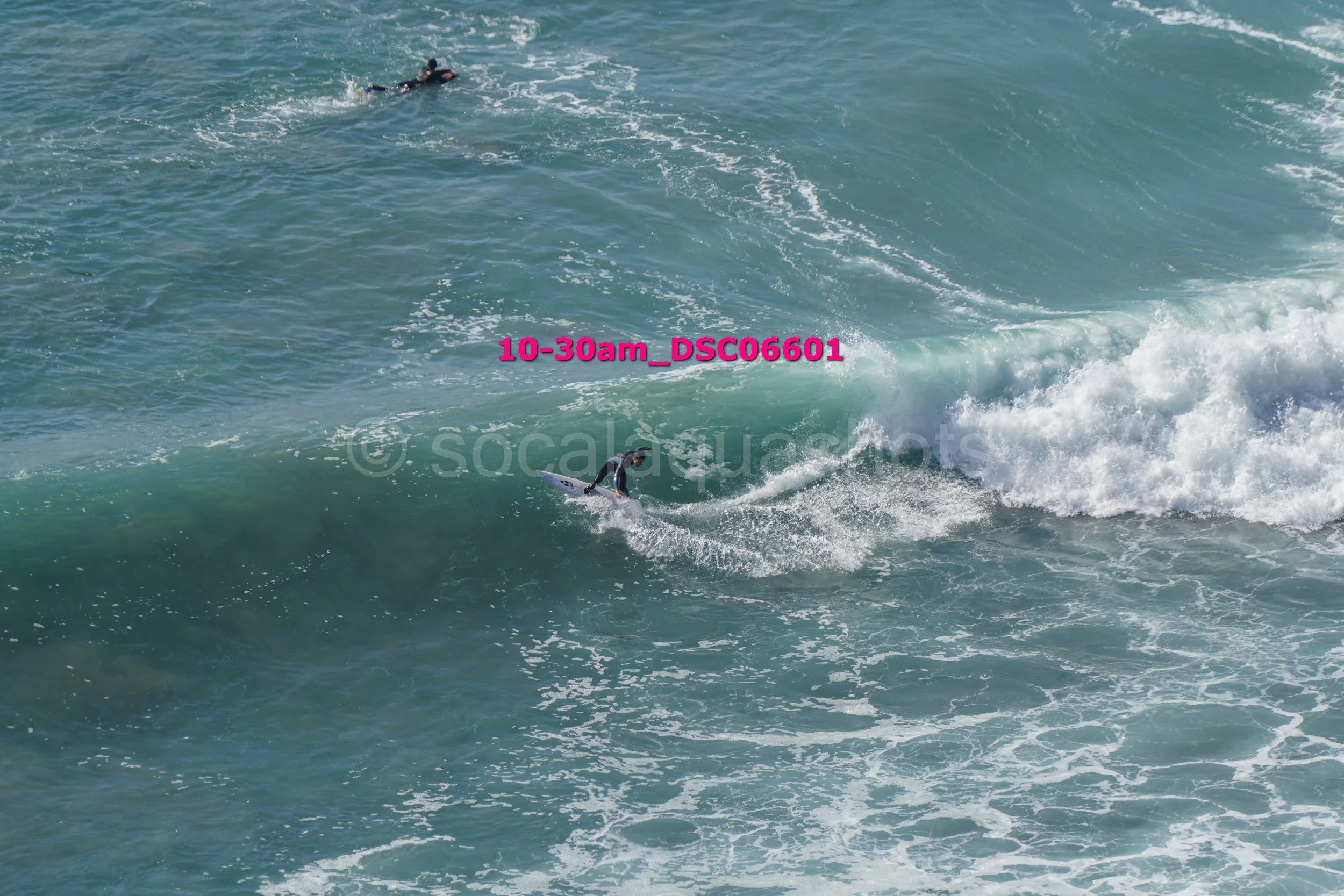 A person surfing on a wave in the ocean with another surfer or swimmer further in the background.