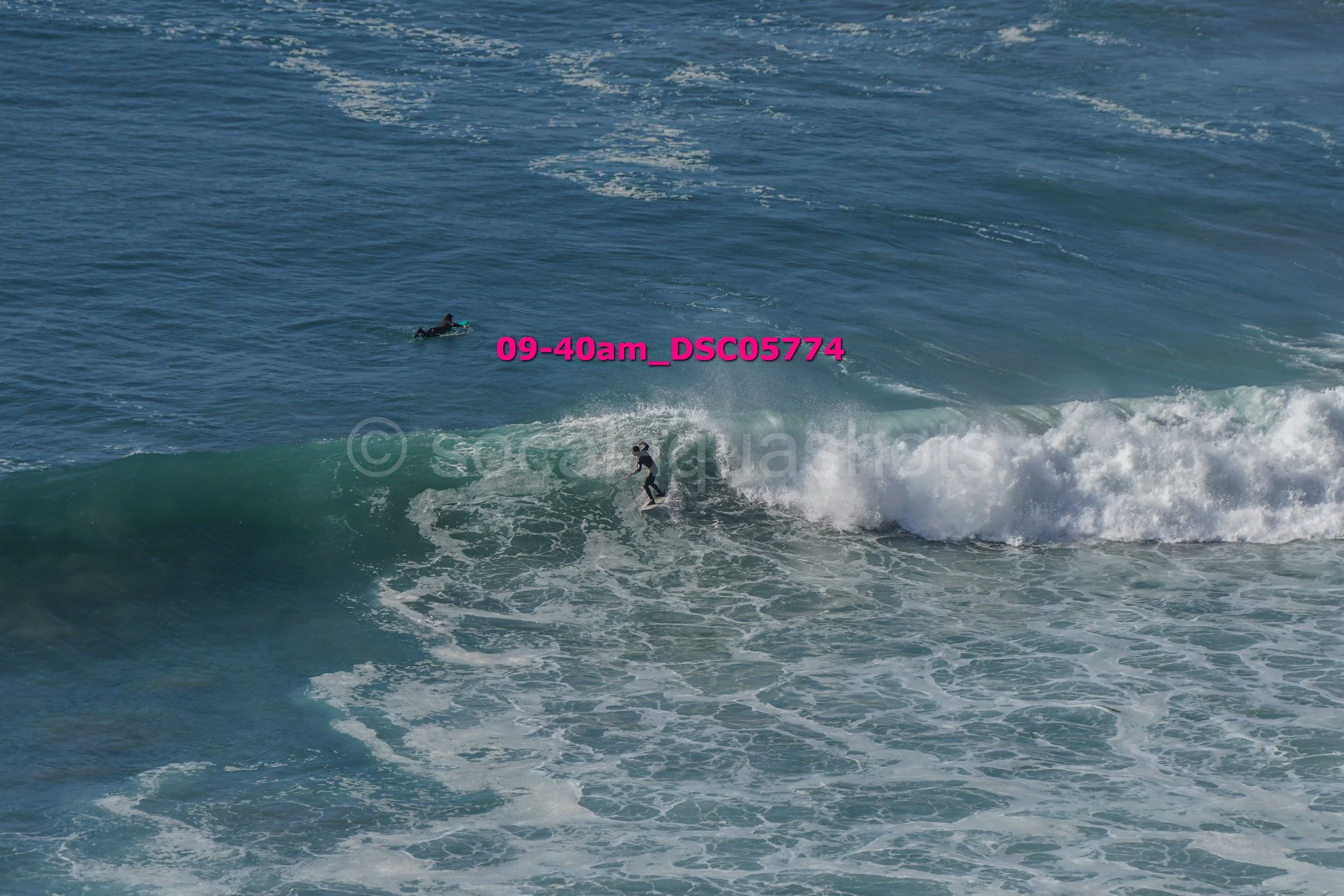 A person surfing on a wave in the ocean, with another person lying on a surfboard in the water nearby.