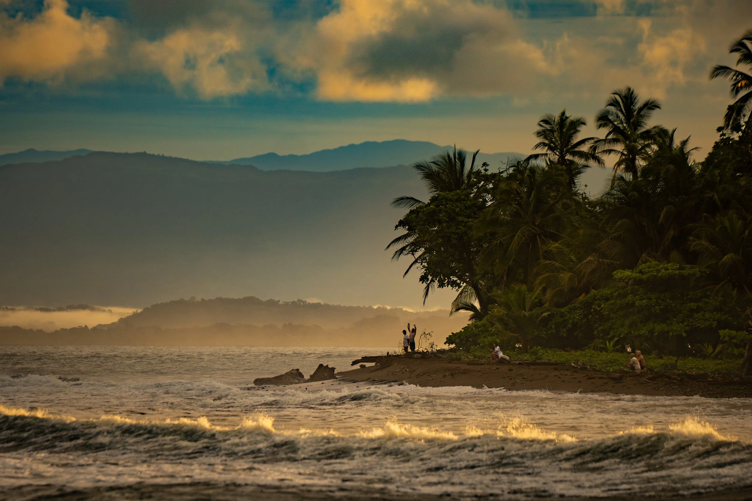 Sunset over a tropical beach with palm trees, mountains in the background, and a few people near the water and on the shore.