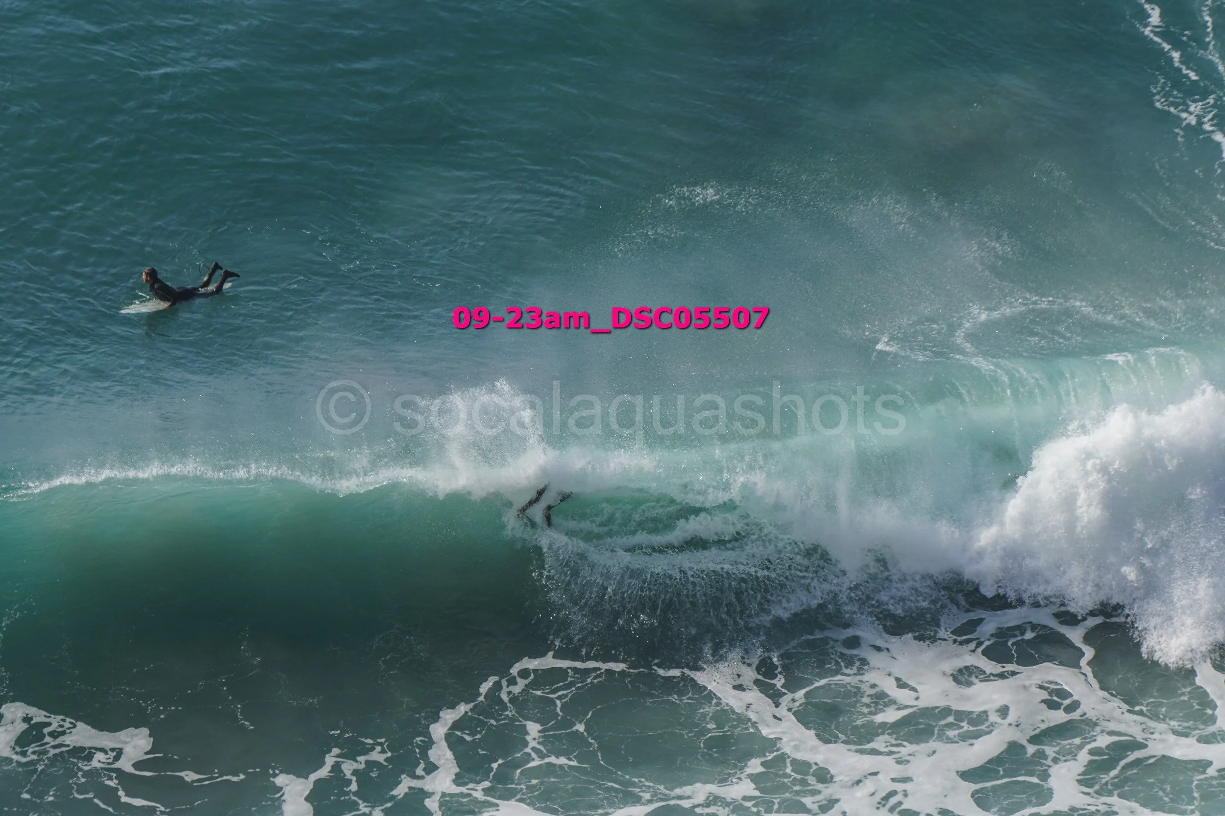 A person surfing on a large ocean wave with another person swimming nearby.