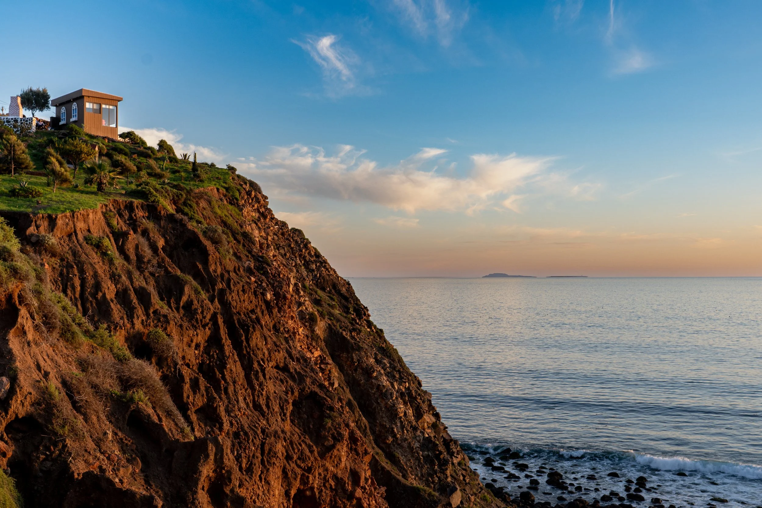 A picturesque view of a coastal cliff with a house on top, overlooking the ocean during sunset with a partly cloudy sky.