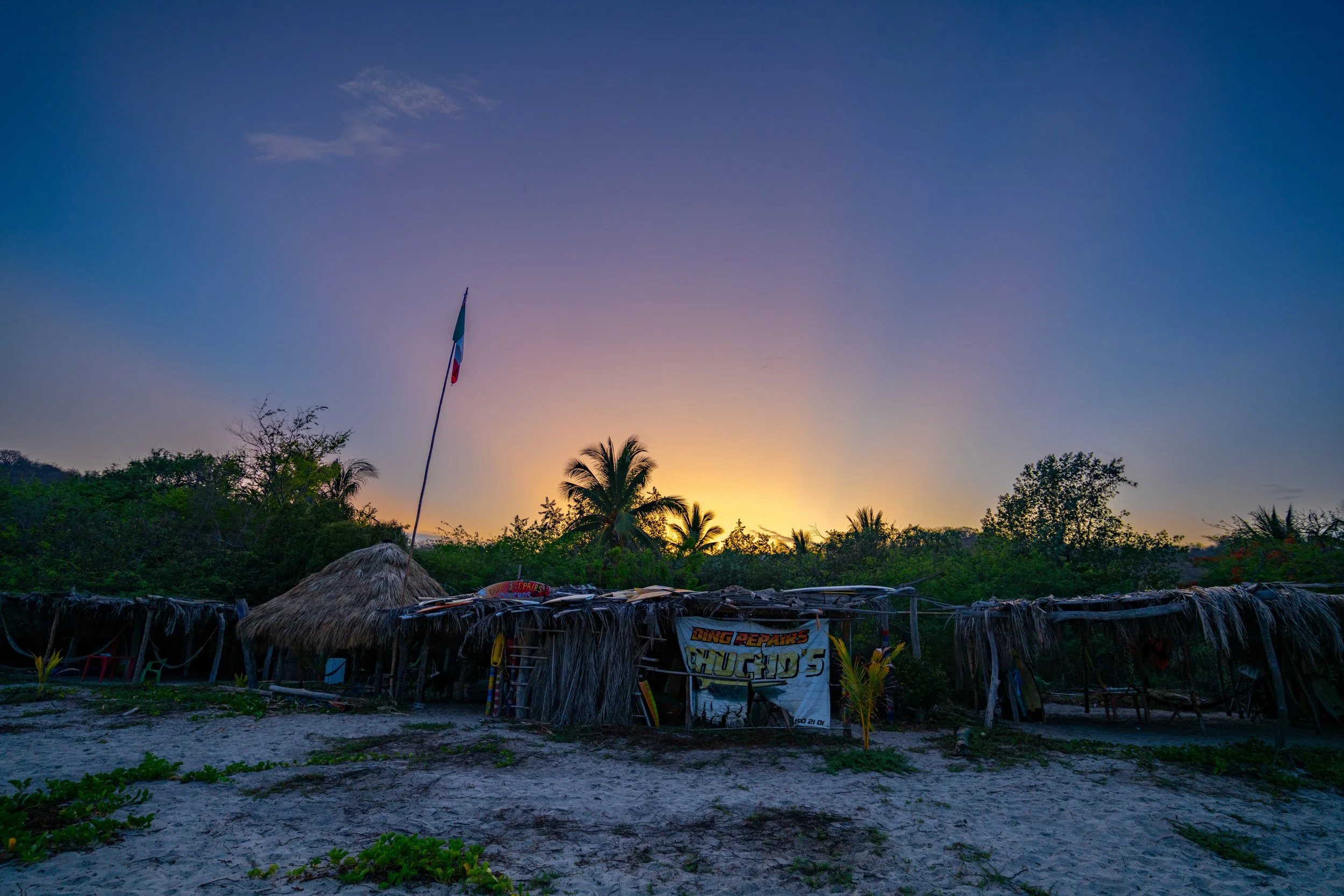 Sunset over a tropical village with palm trees, thatched roofs, and a flagpole with a flag, surrounded by lush greenery and sandy ground.
