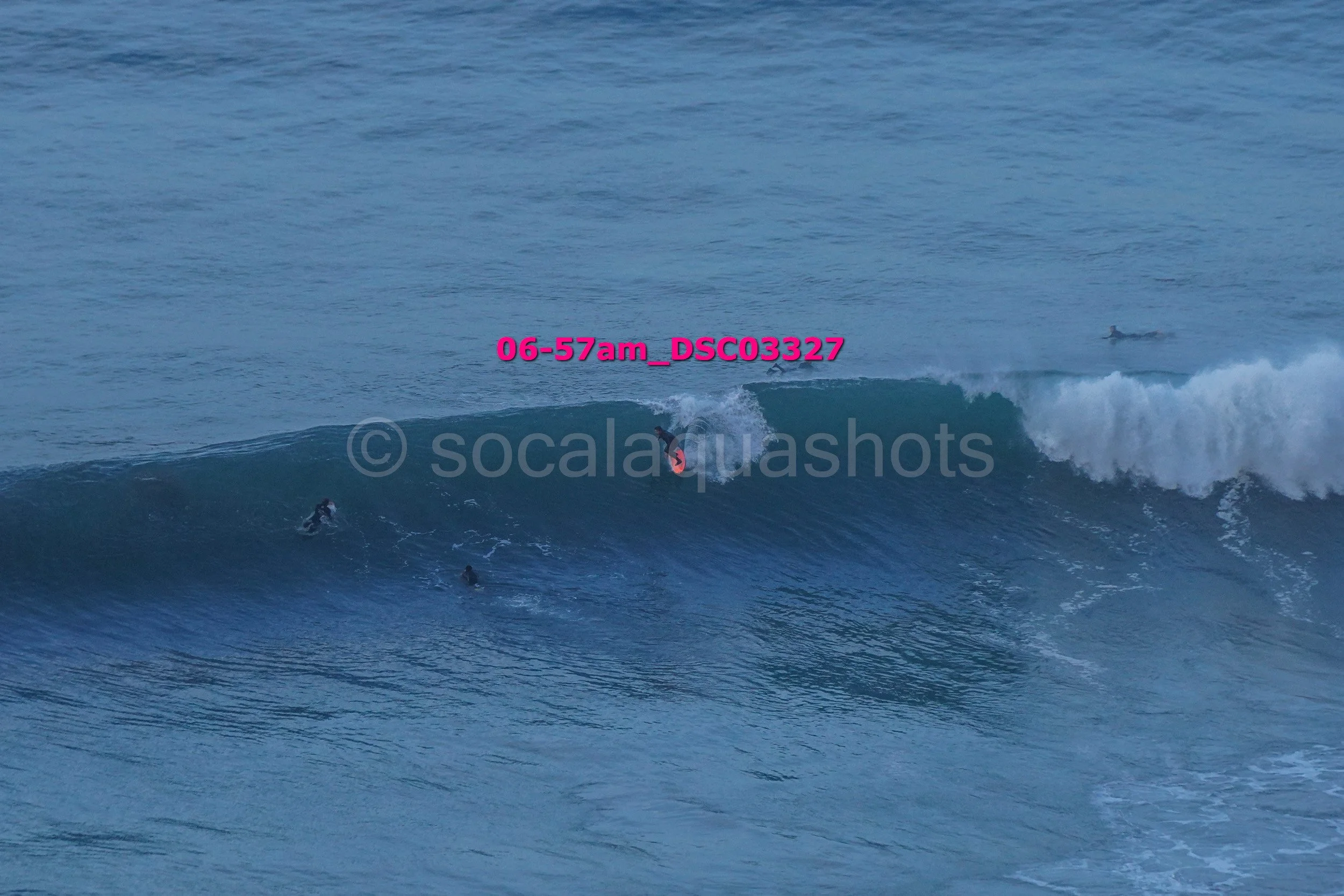 Surfers riding a large wave in the ocean, with some surfers in the water and one surfer on a wave near the shoreline.