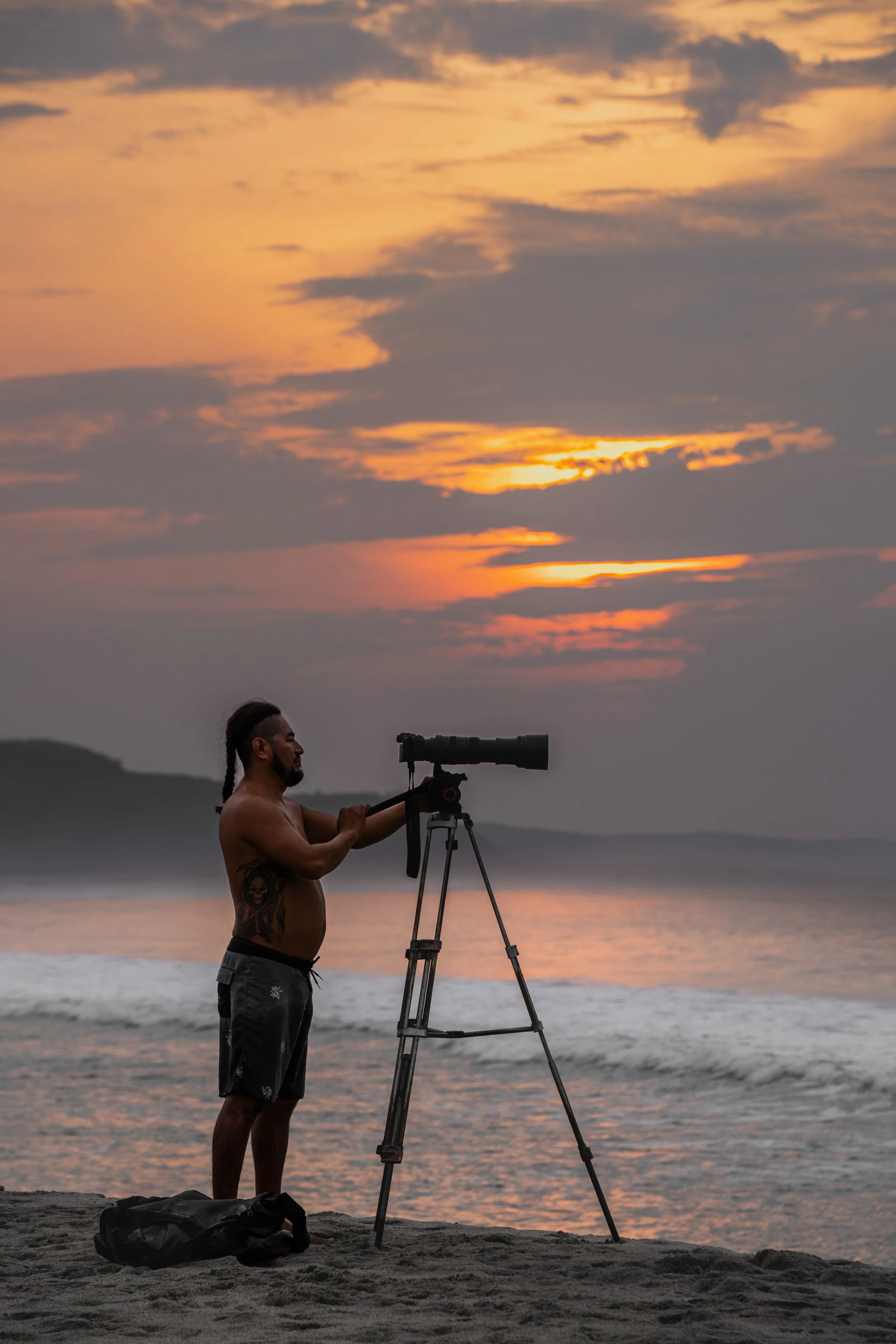 A shirtless man with tattoos on his left arm and chest stands on a sandy beach at sunset, adjusting a camera on a tripod. The sky is filled with orange, yellow, and gray clouds over the water, with distant landforms visible in the background.