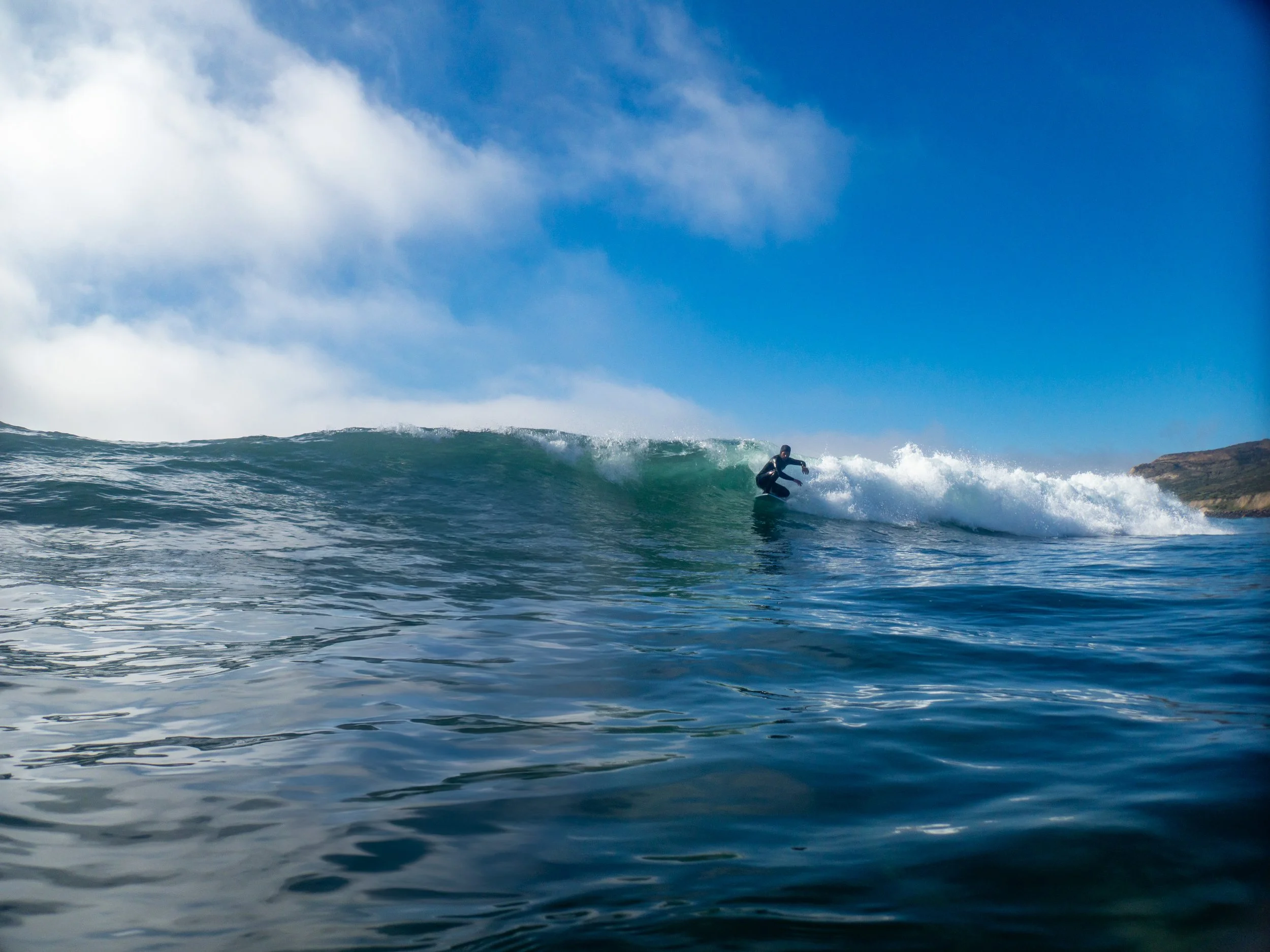 A person surfing on a wave in the ocean under a blue sky with some clouds.