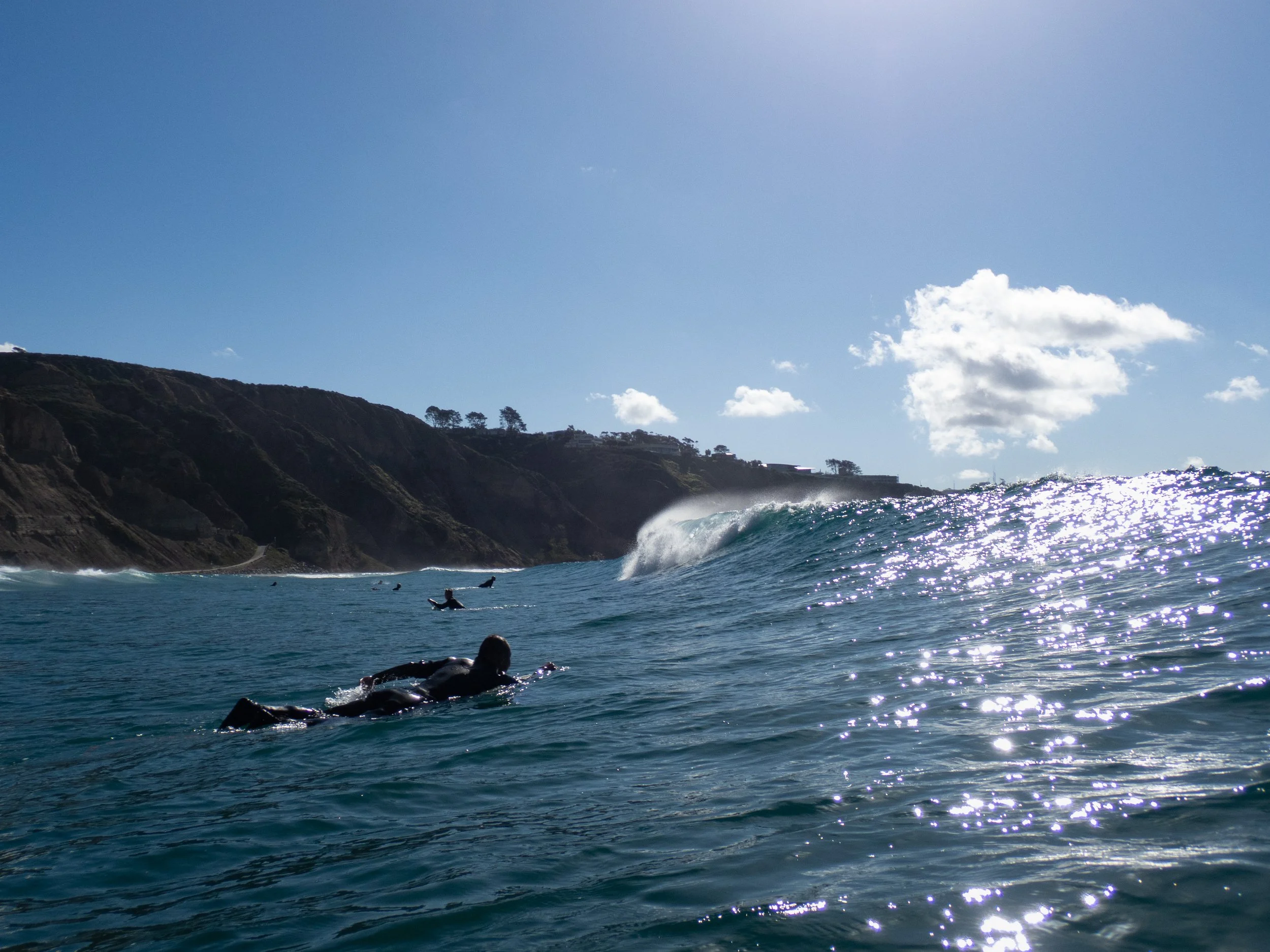 People surfing in the ocean near a rocky coastline on a sunny day with a few clouds in the sky.