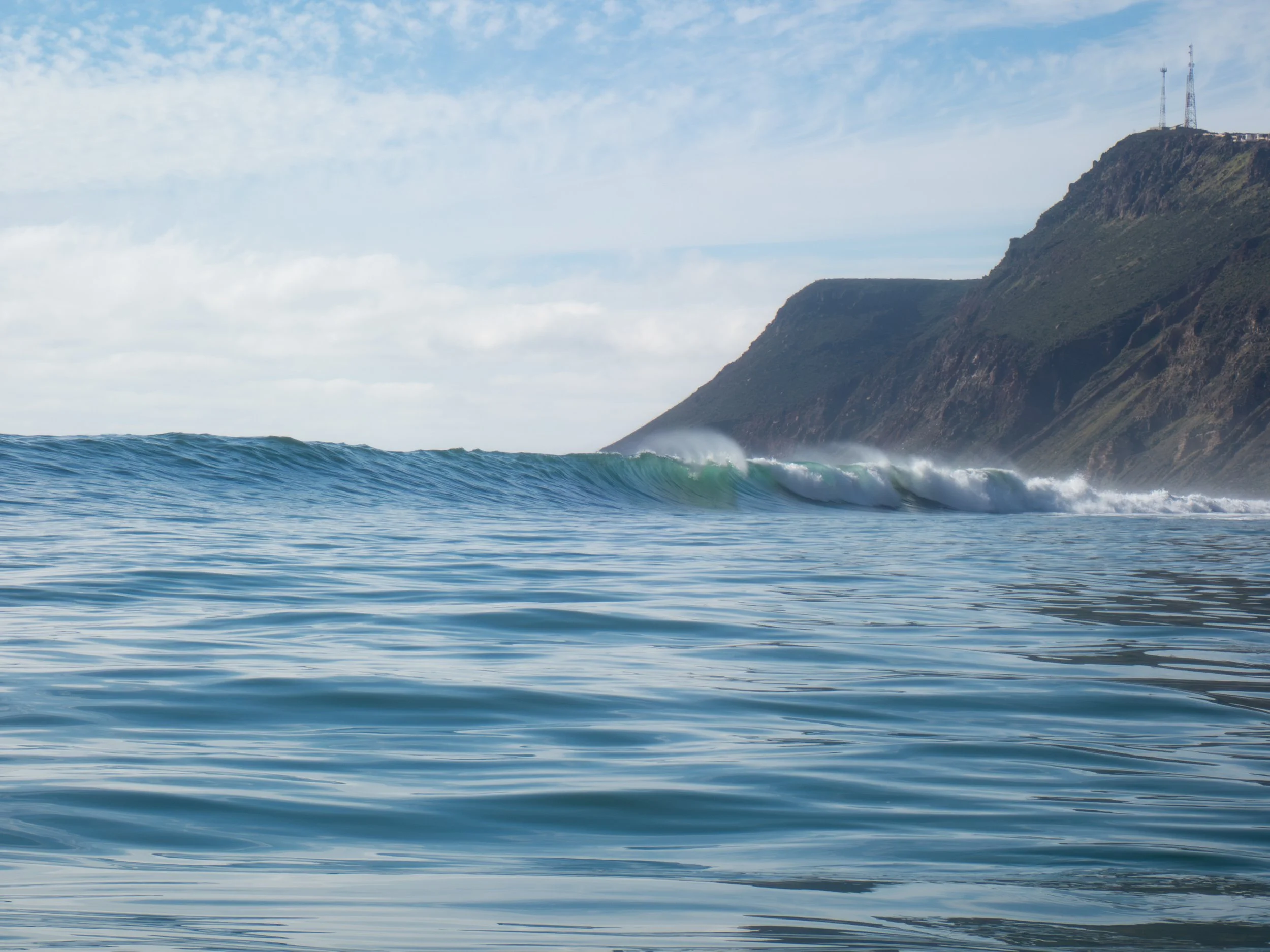 Ocean waves near the shoreline with a mountain and communication towers in the background under a partly cloudy sky.