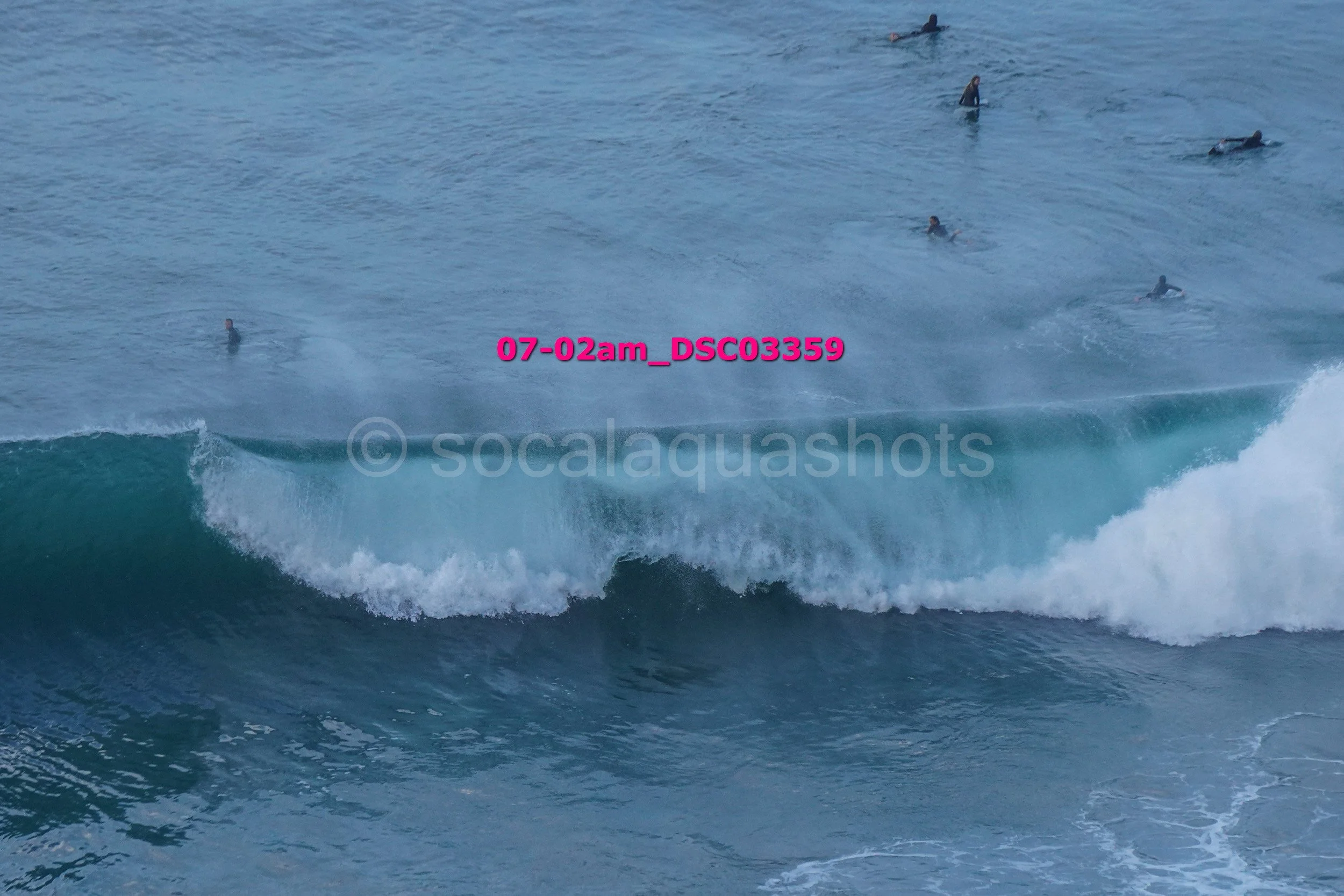 Several surfers paddling in the ocean with a large breaking wave in the foreground