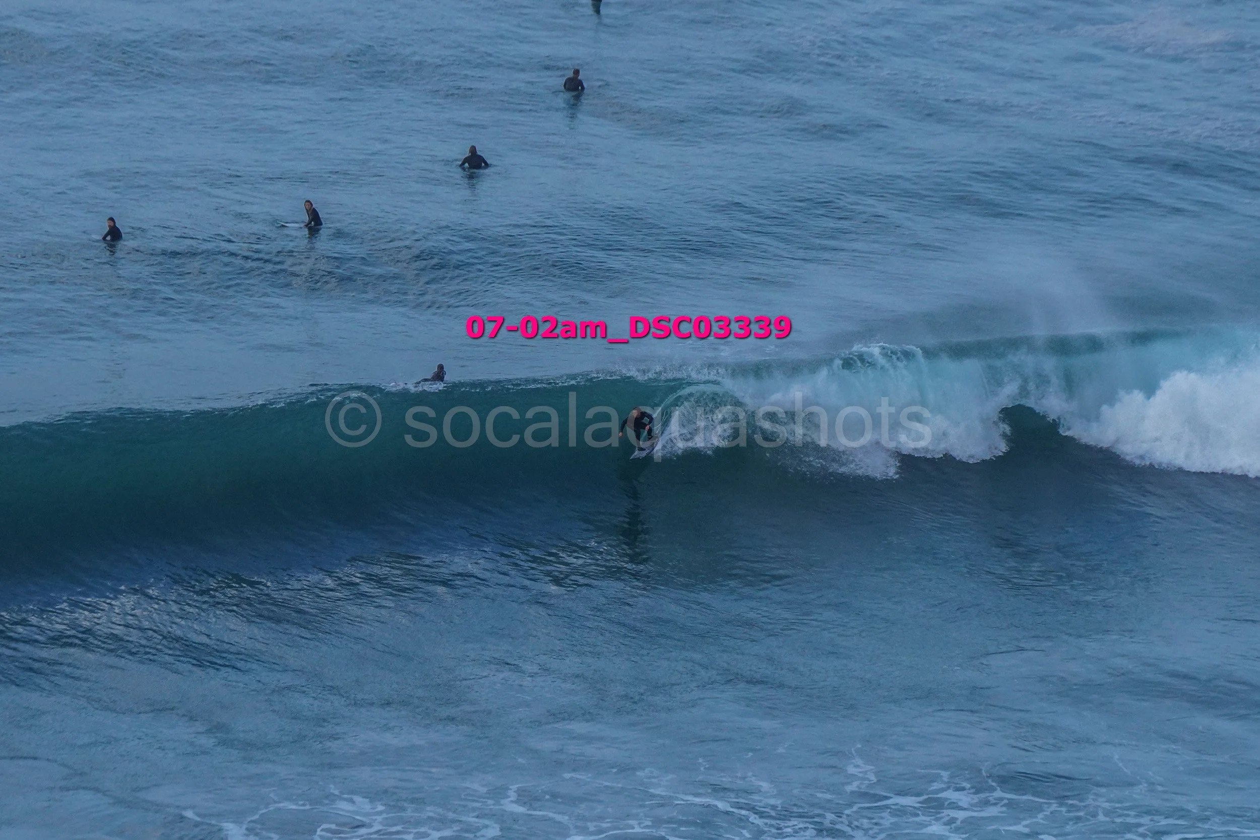 A surfer riding a wave with several surfers and paddleboarders in the water nearby, overcast sky.