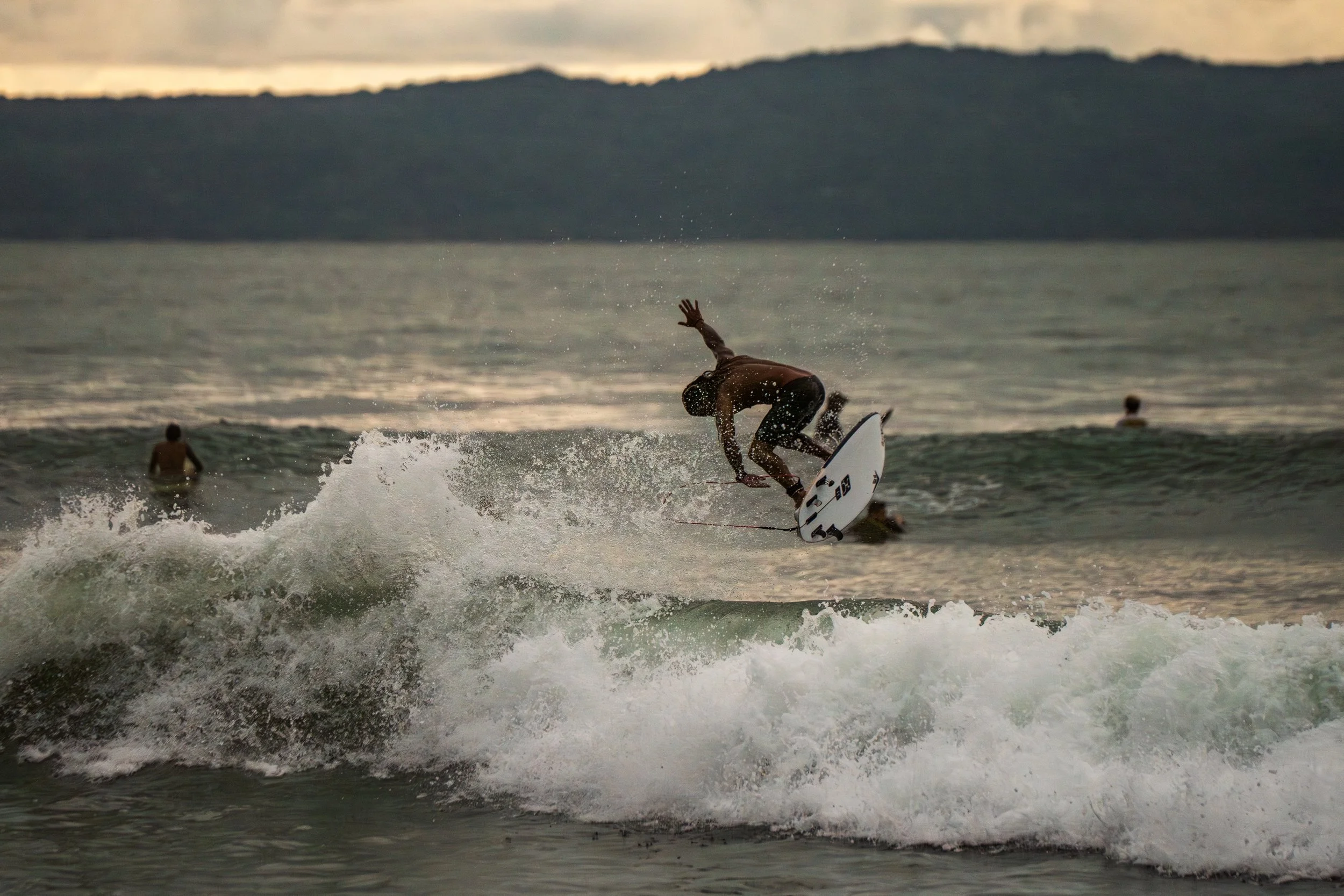 A person surfing on a wave in the ocean during sunset or sunrise with a few other people in the water and a hilly or mountainous landscape in the background.