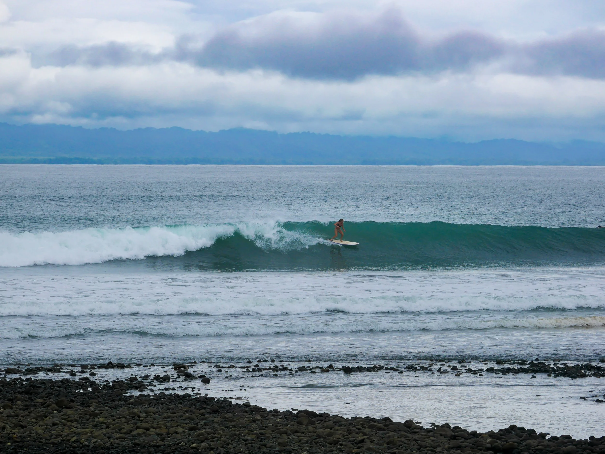 Surfer on a wave in the ocean with a rocky shore in the foreground and cloudy sky above.