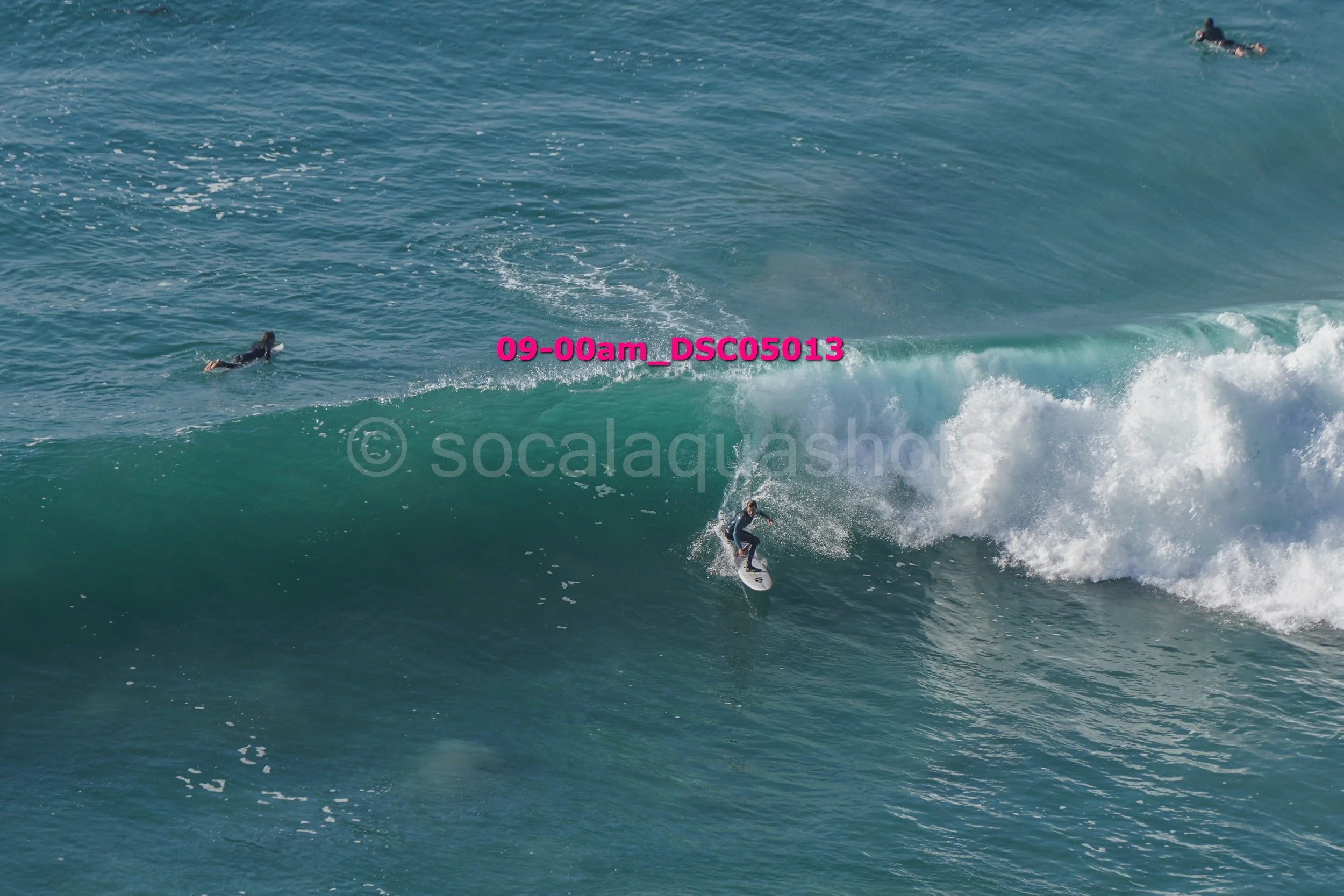 A person surfing on a large ocean wave with two other surfers visible in the water nearby.
