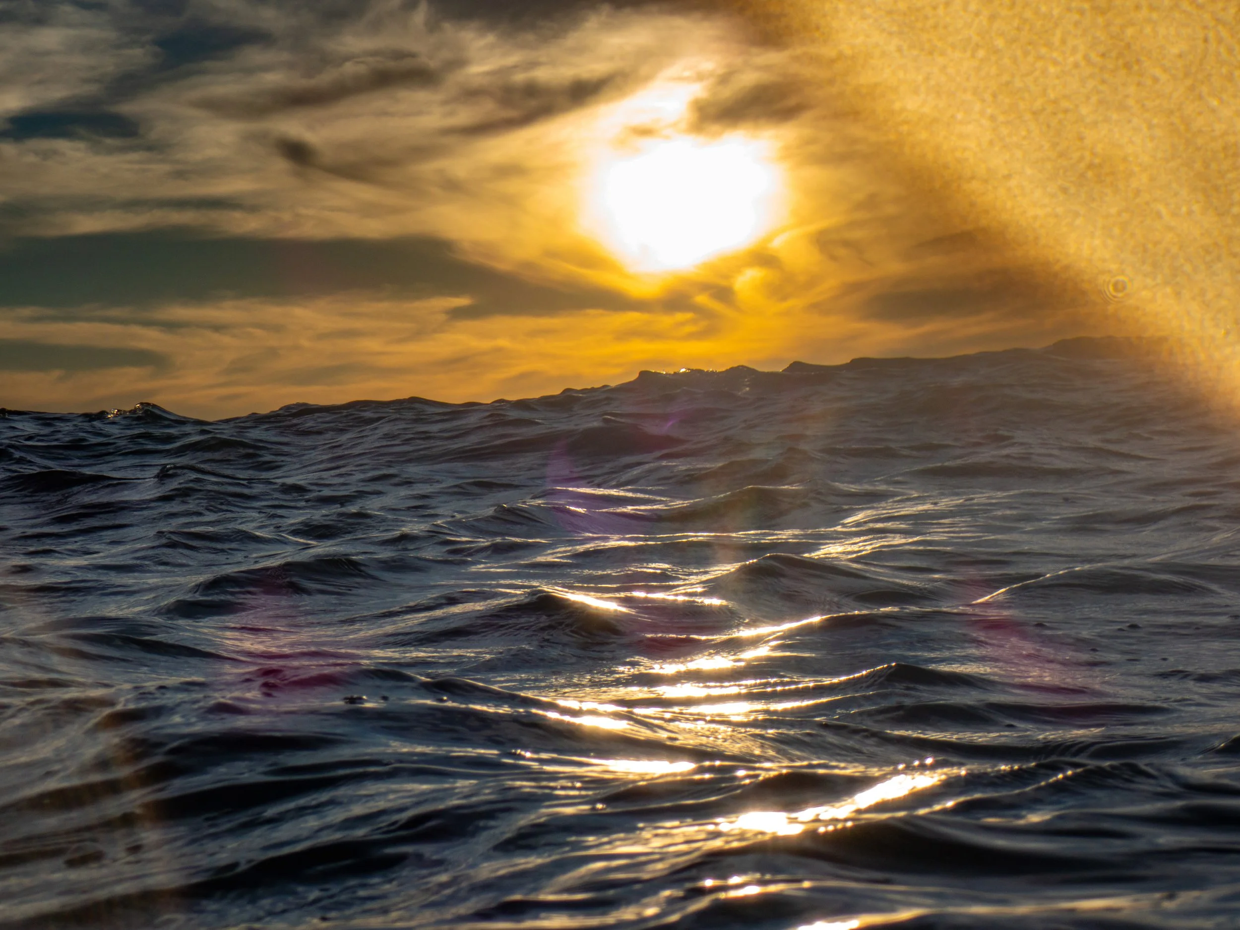 Ocean waves at sunset with the sun partially obscured by clouds in the sky.