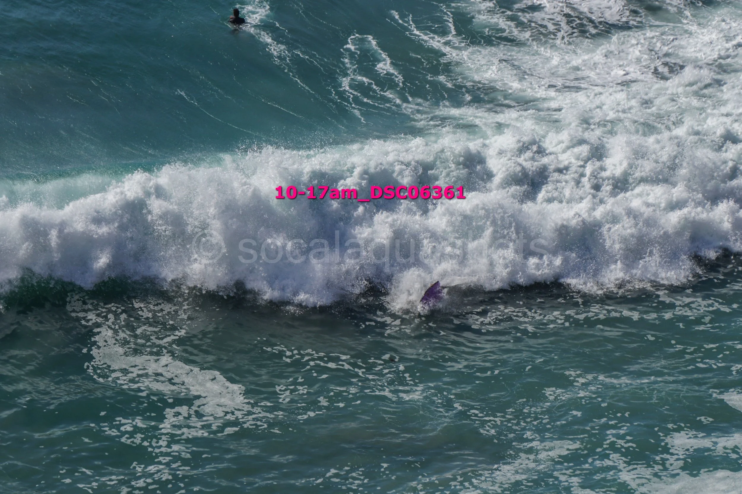 A surfer's purple surfboard is partially submerged in the ocean waves, with a person seen in the water in the background.