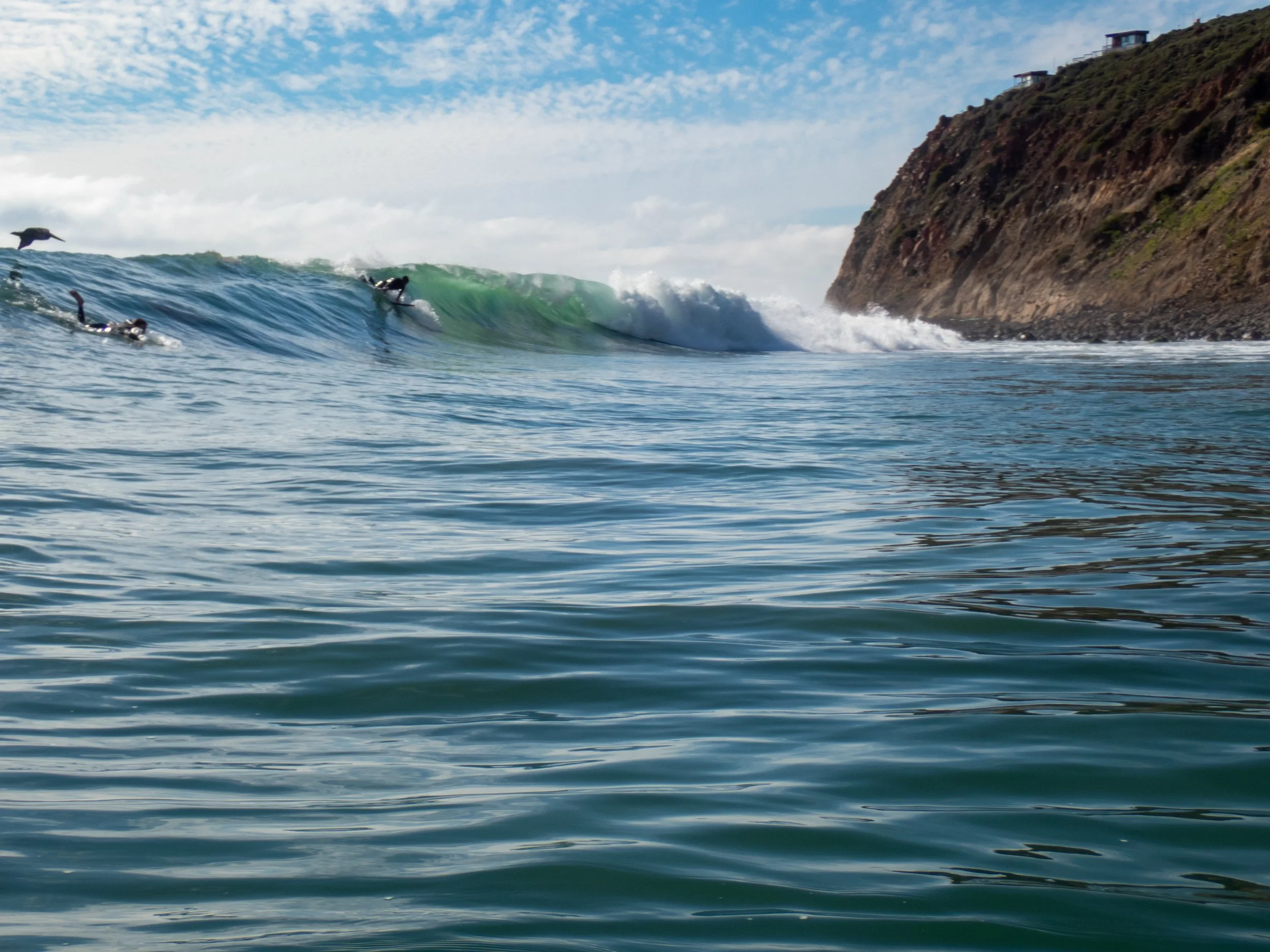 Surfers riding waves near a rocky coastline under a partly cloudy sky.