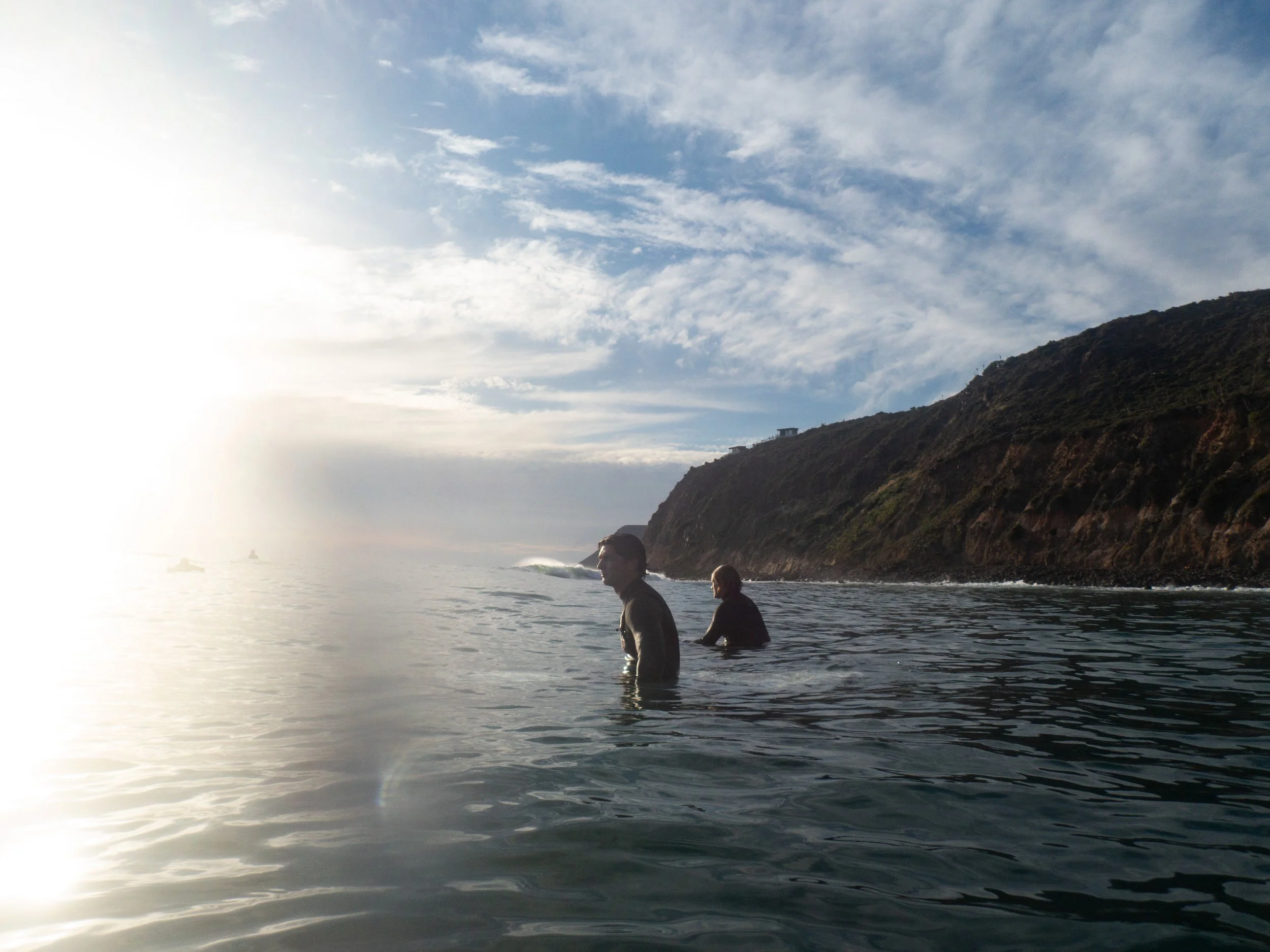 Two men are standing in the ocean near a rocky coastline during sunset, with partly cloudy sky above.