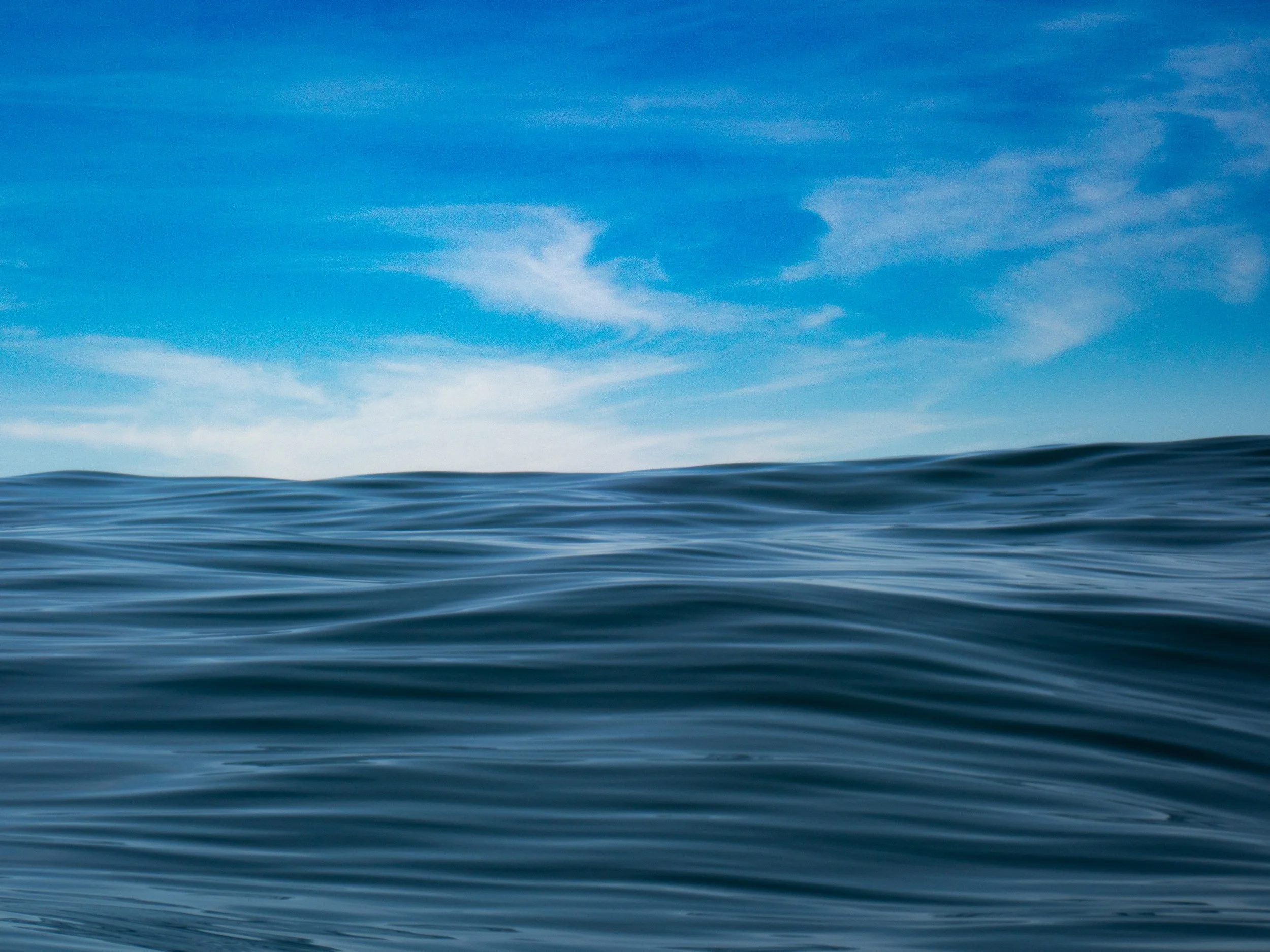 Vast expanse of ocean water with gentle waves, under a clear blue sky with wispy clouds.