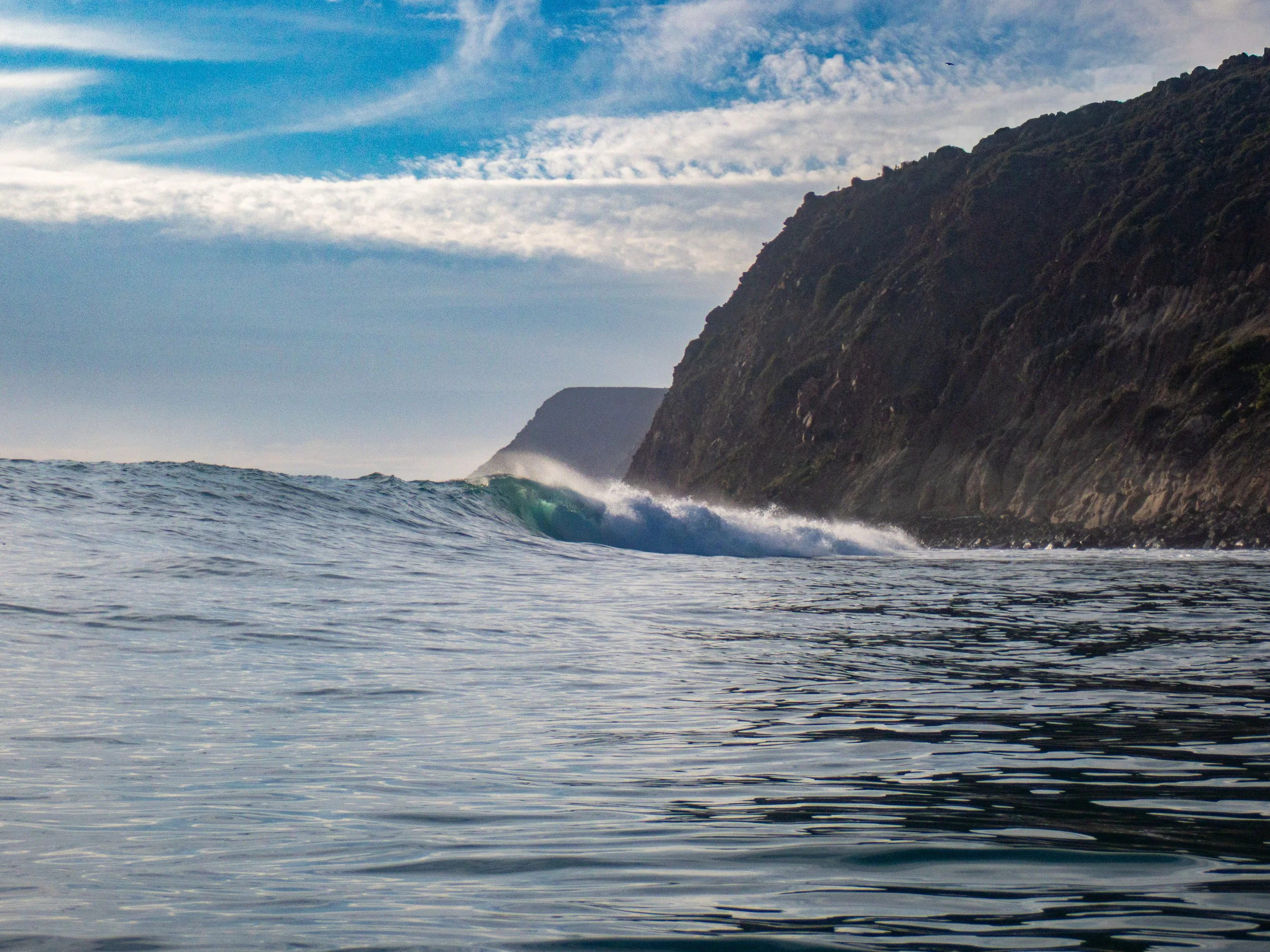 Ocean waves near a rocky coastline with cliffs and a partly cloudy sky.