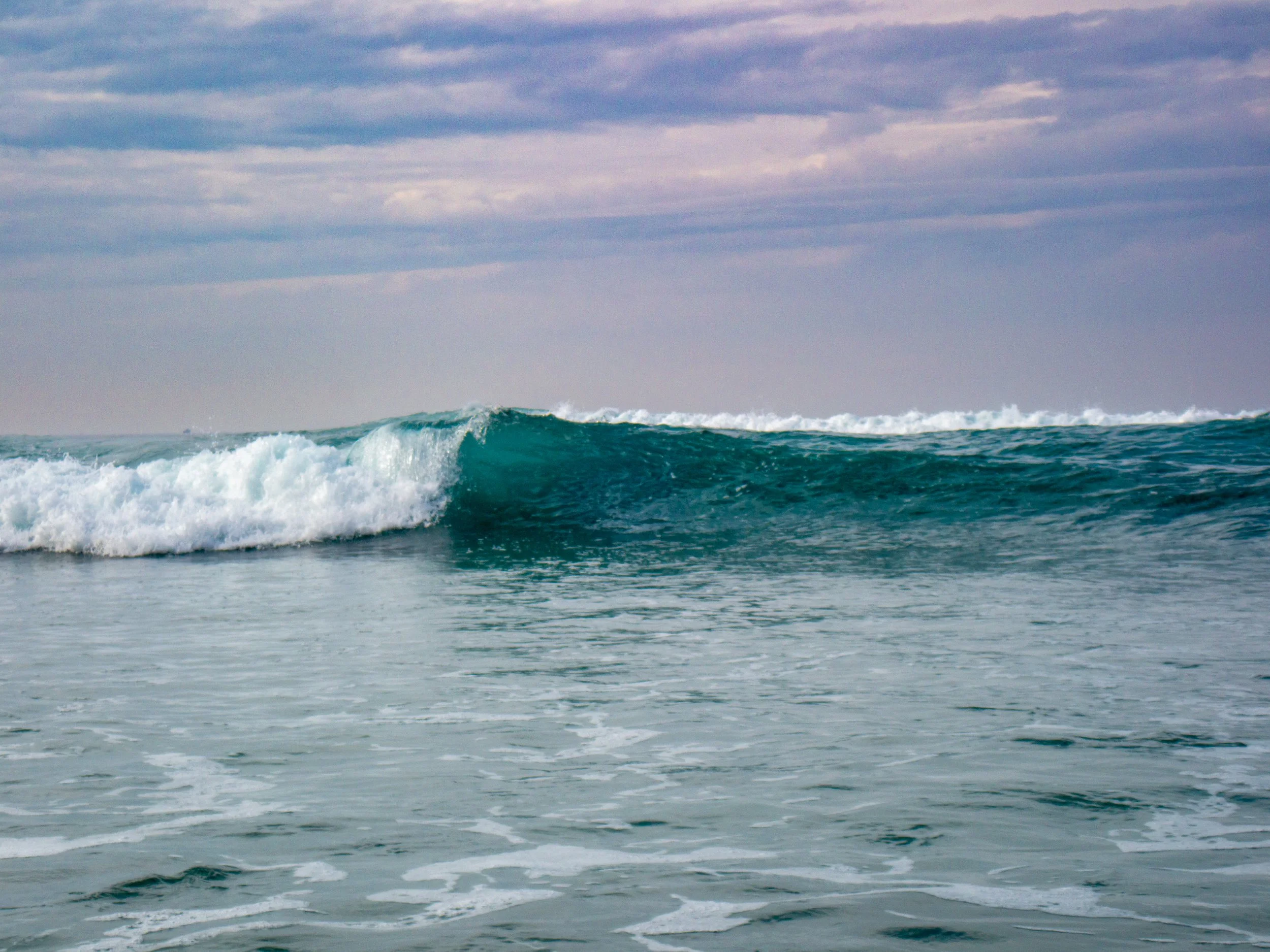 A calm ocean with a single breaking wave under a cloudy sky.