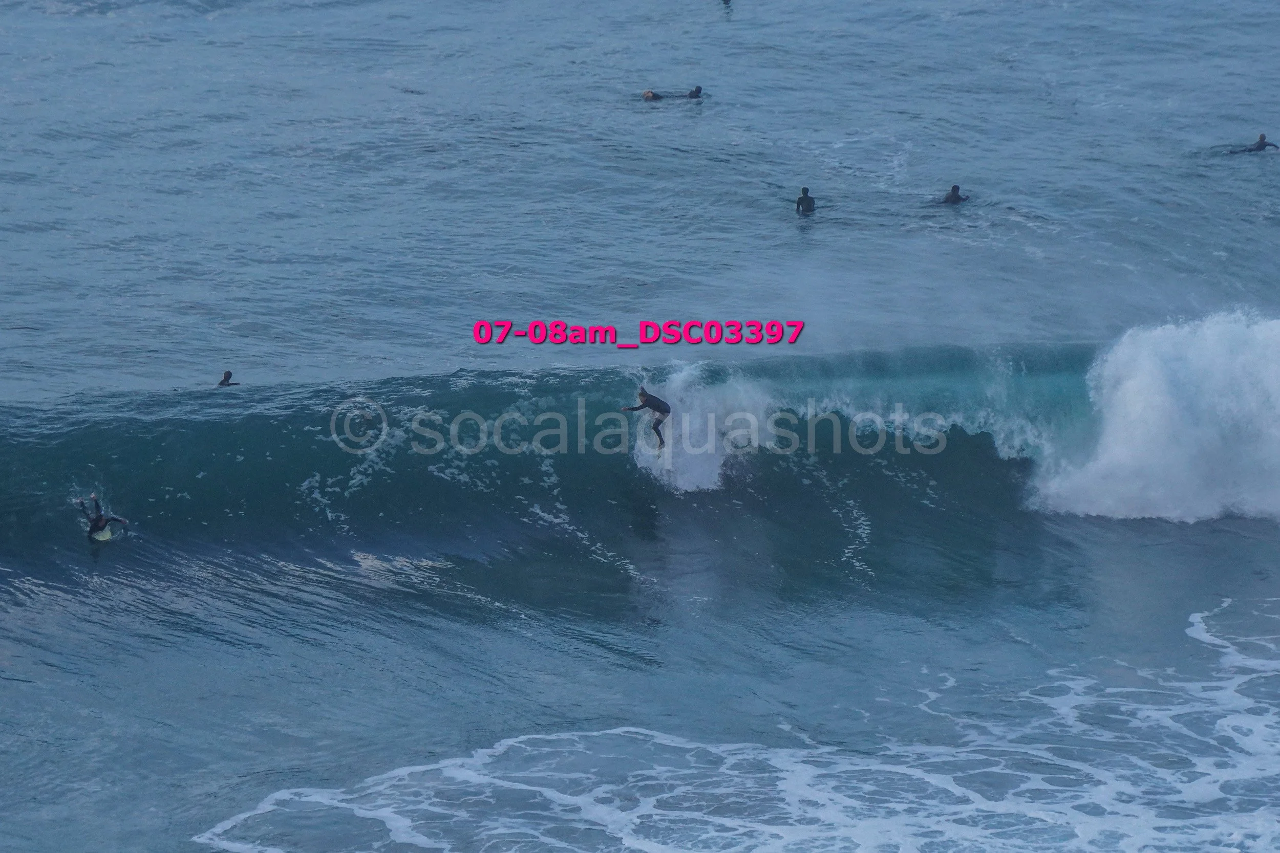 Surfer riding a large wave with several people swimming and surfing in the background in the ocean.
