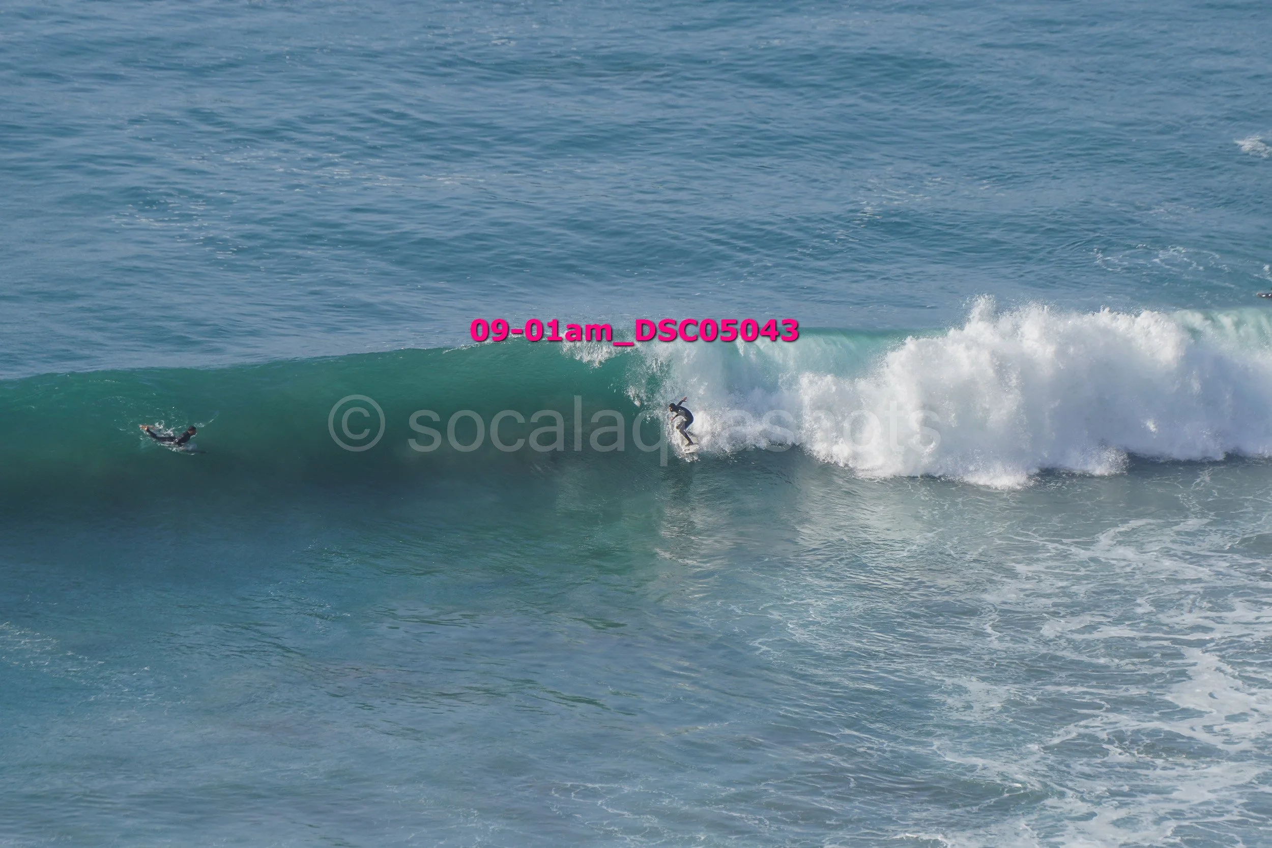 Surfer riding a wave with a second surfer paddling nearby in the ocean.