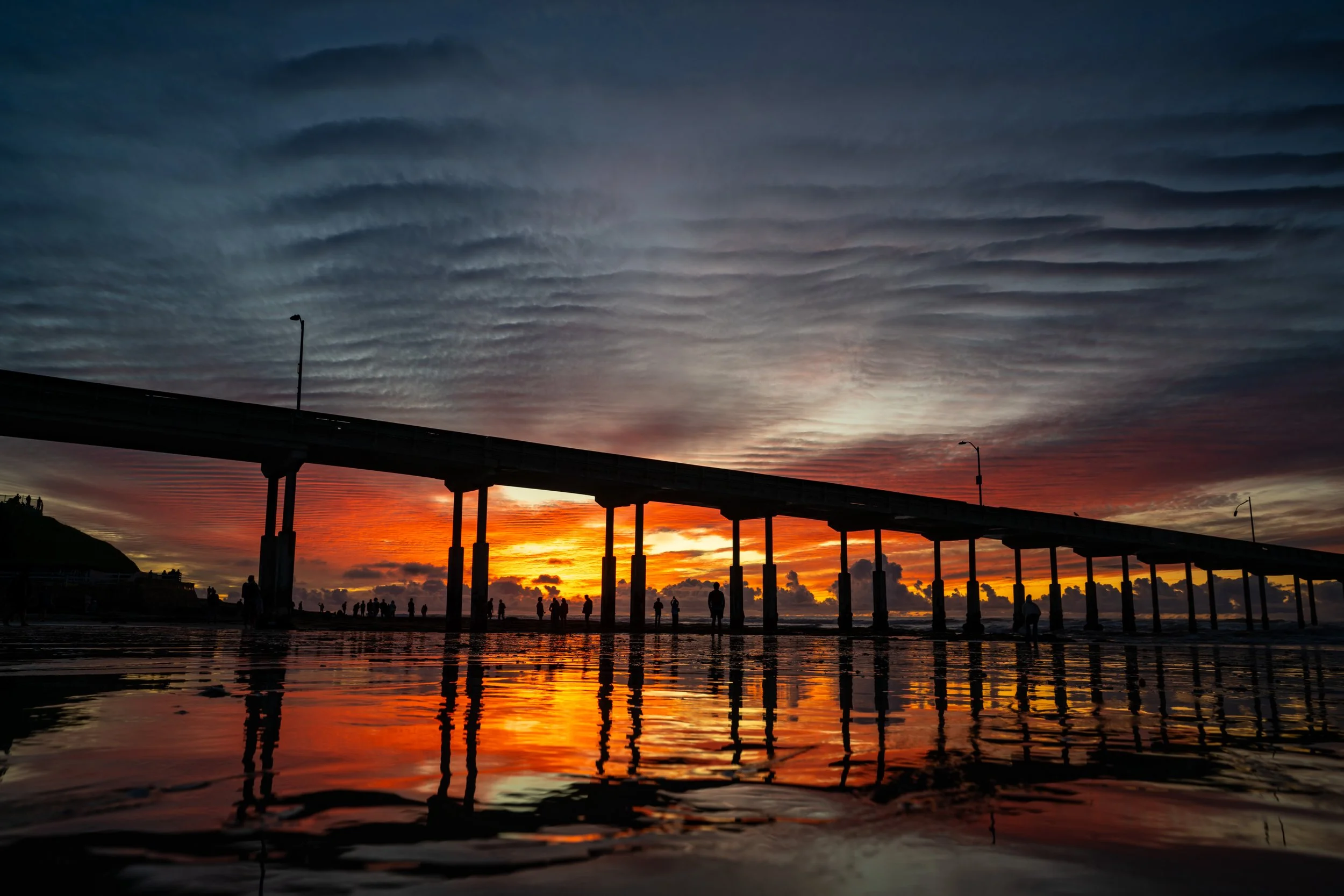 Silhouettes of people walking on a beach at sunset, with a long pier extending into the water. The sky is painted with vibrant orange, red, and purple hues, and the water reflects these colors. Cloud formations create a dramatic backdrop.