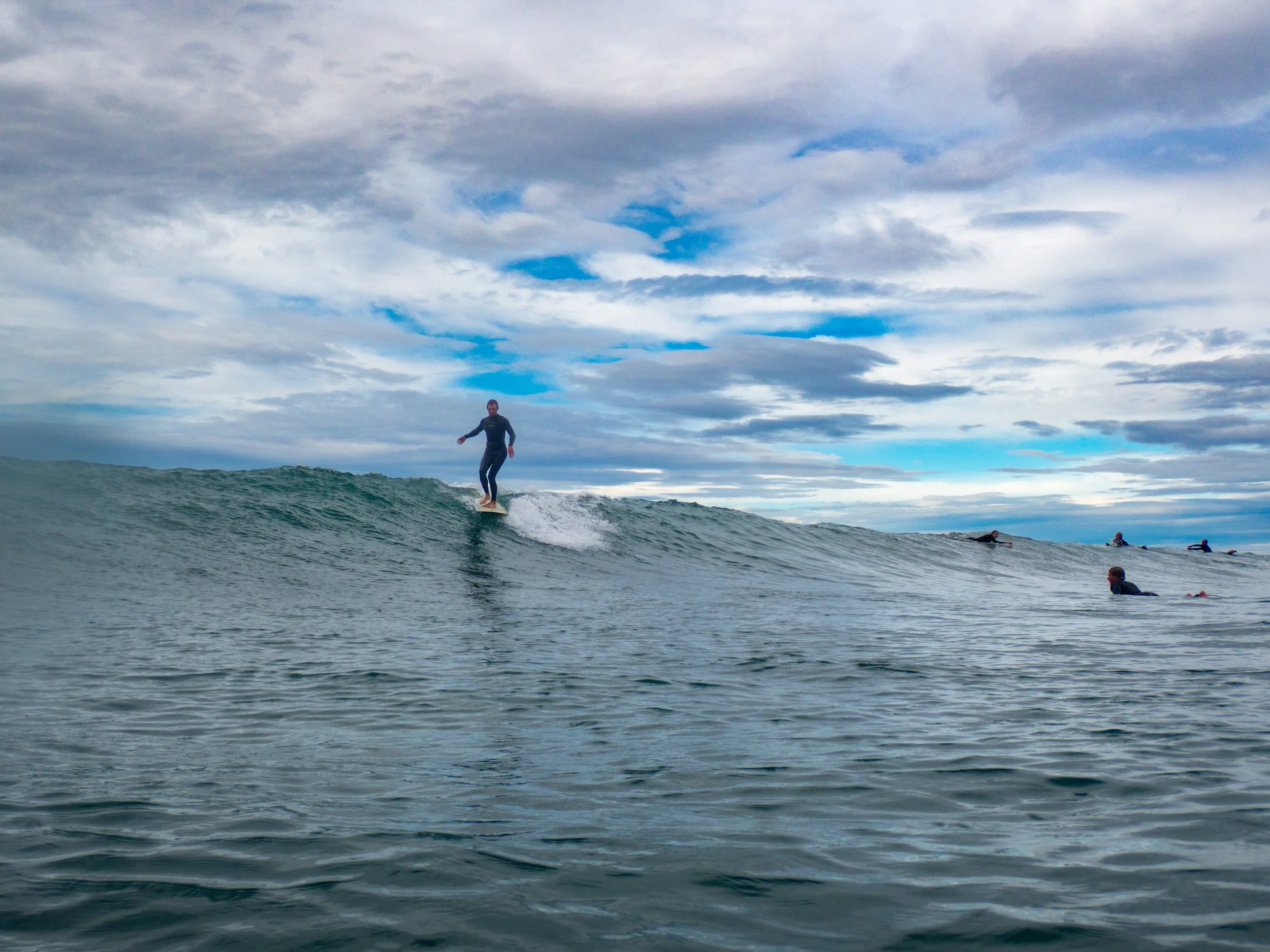 A person surfing on a wave with several other surfers in the water under a cloudy sky.