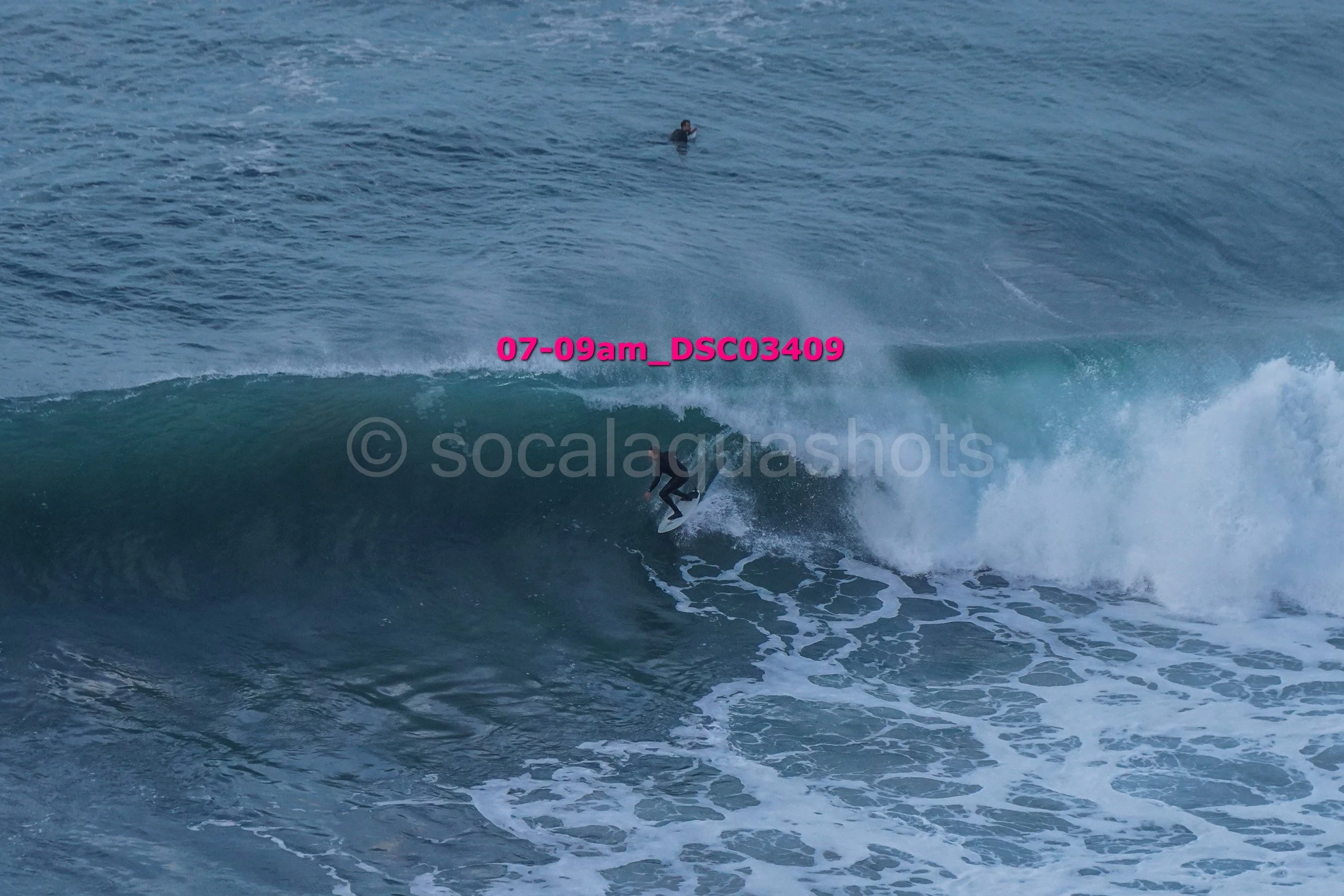 A person surfing on a wave in the ocean, with another person swimming in the background.