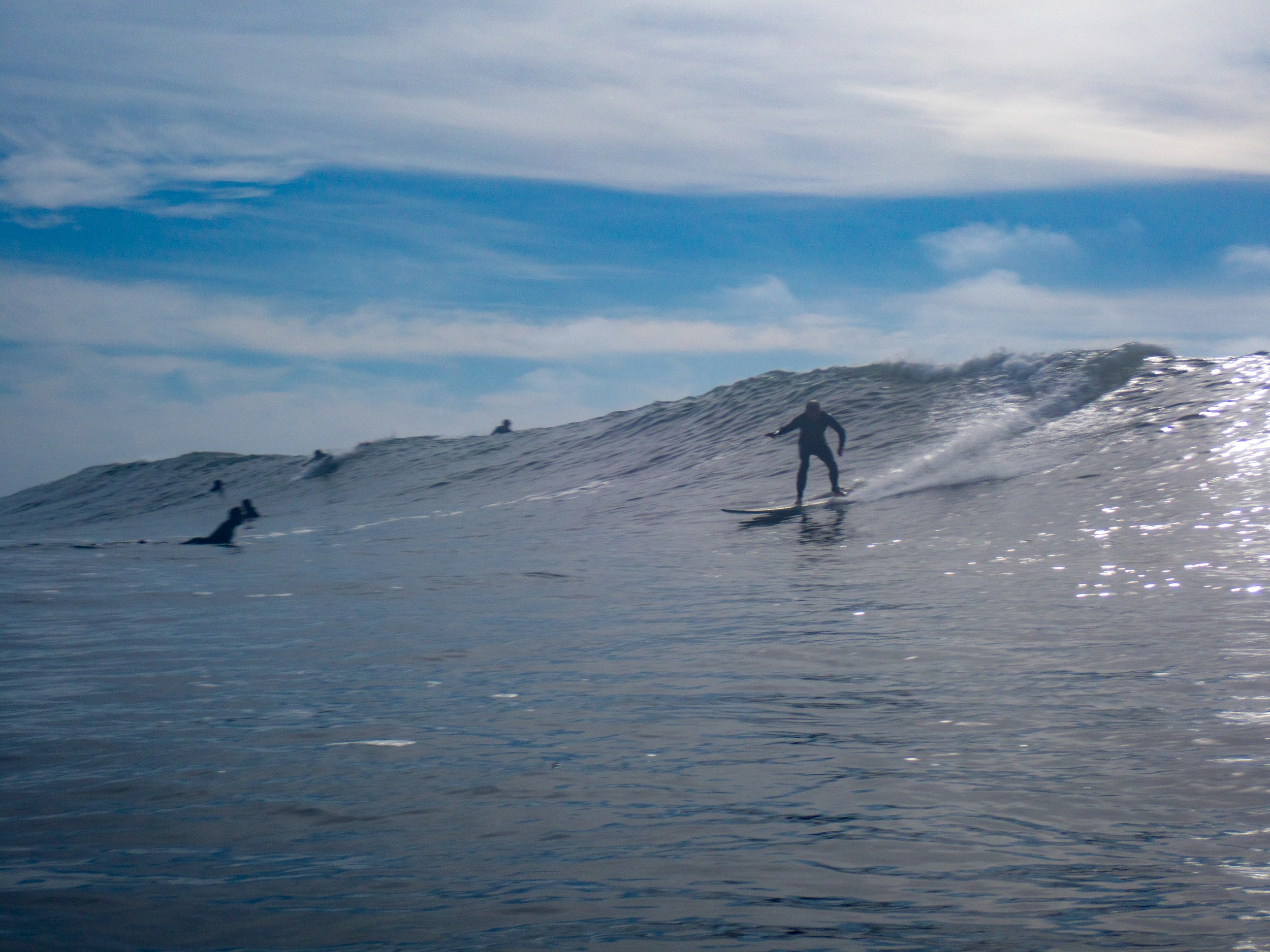 Surfer riding a wave with several other surfers in the water under a partly cloudy sky.