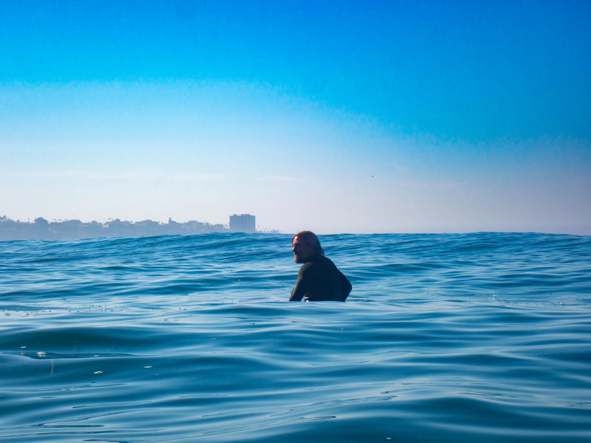 A person with long hair swimming in the ocean with cityscape in the background.