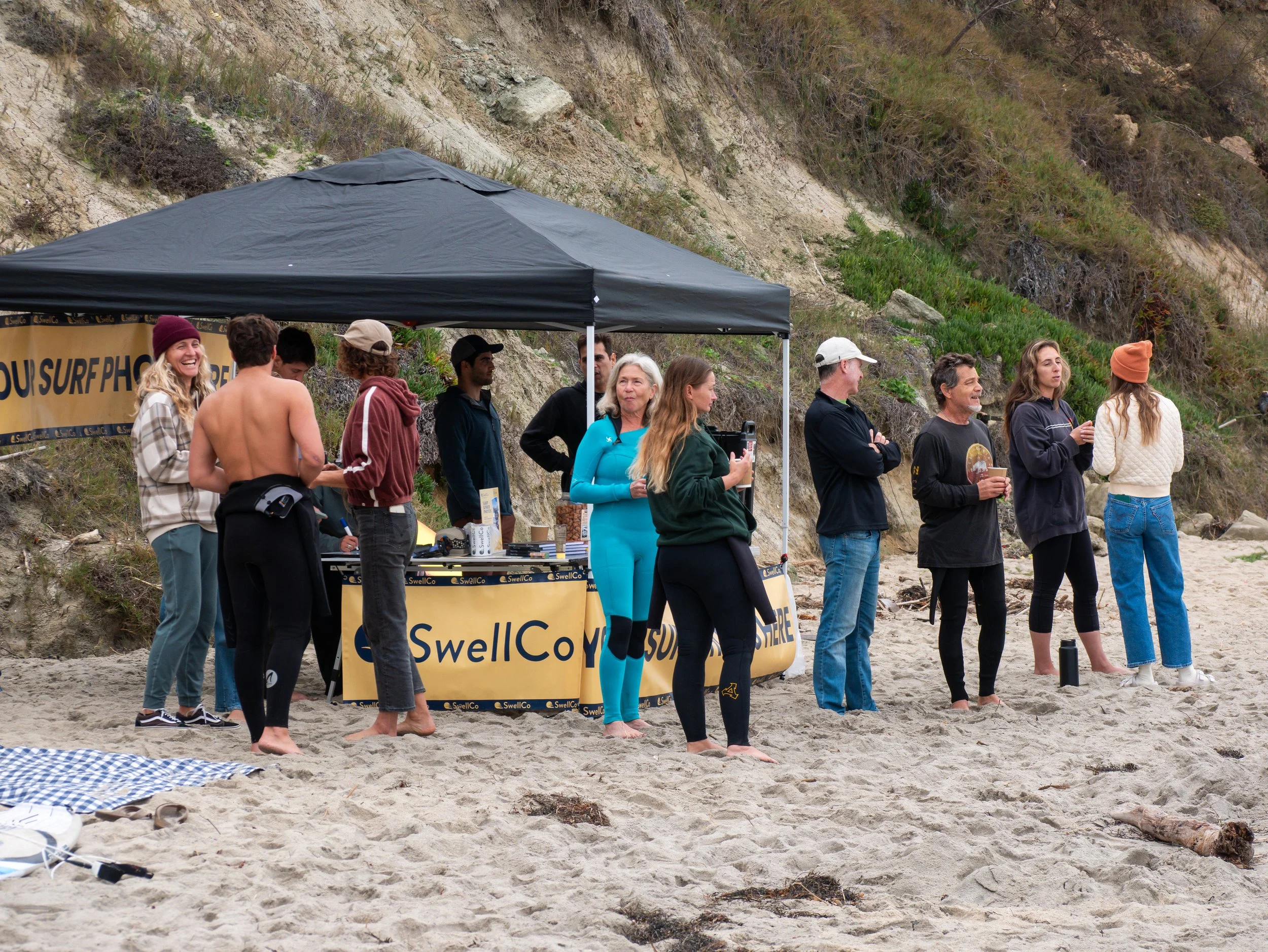 People standing in a line on a beach, some wearing wetsuits, others in casual clothing, with a surf-themed booth and a black canopy in the background.