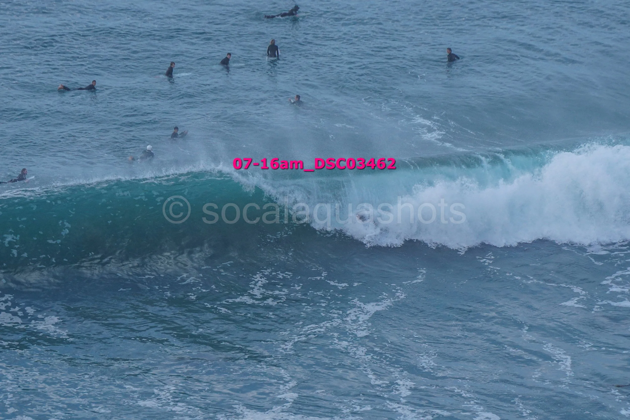 Multiple people in the ocean, some swimming, some surfing near a breaking wave.