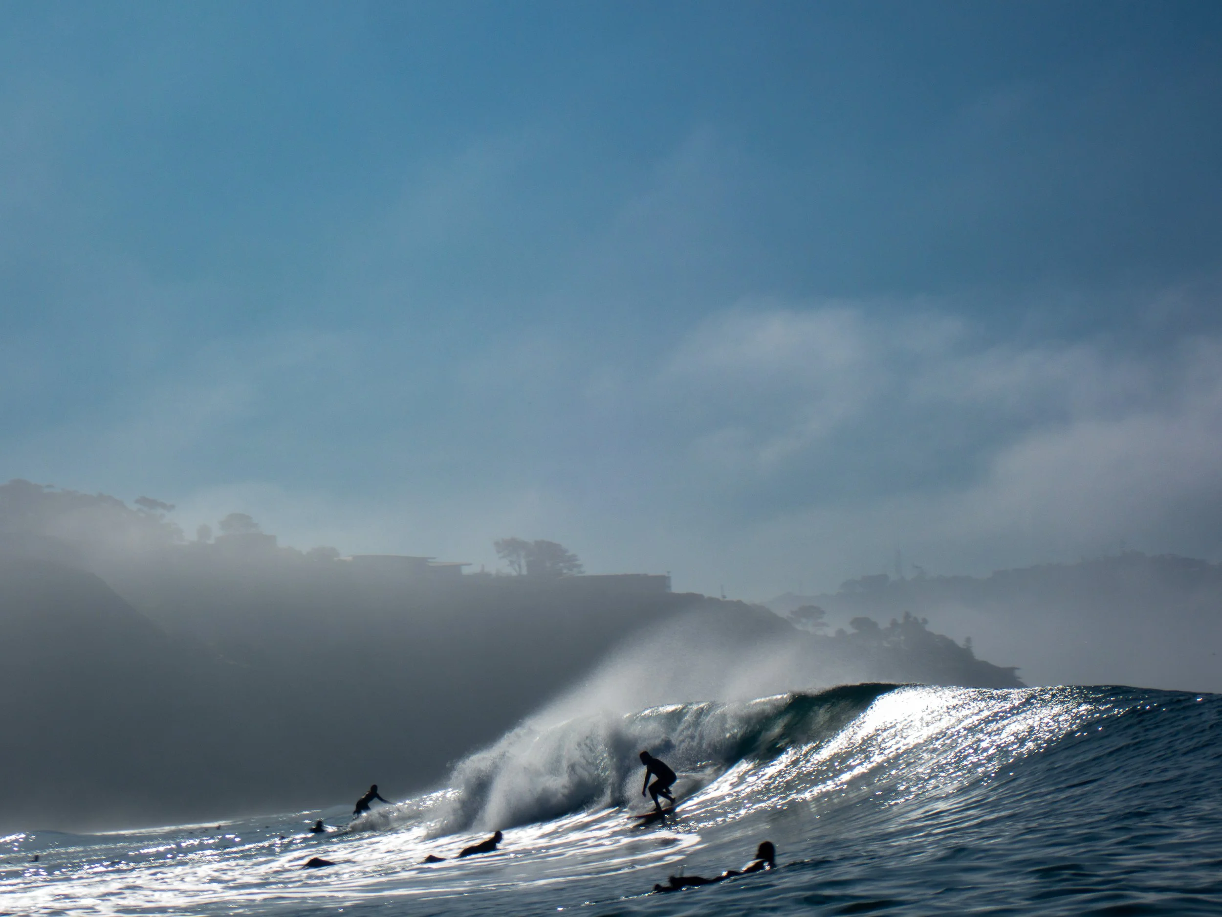 Surfers riding and paddling on ocean waves near the shoreline with misty hills and buildings in the background.