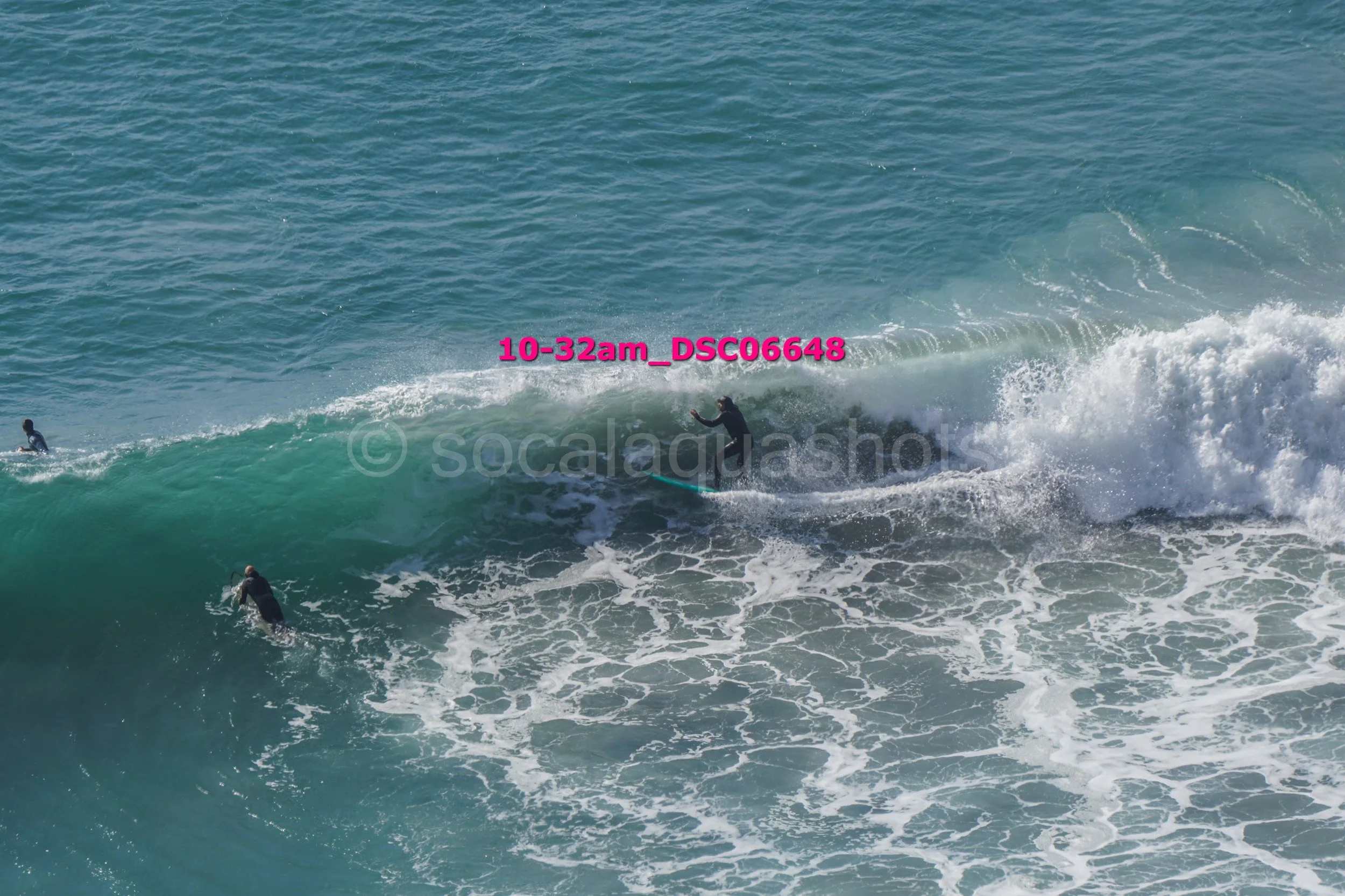 A surfer riding a large wave in the ocean with three other surfers nearby, some in the water and some on their surfboards, under a clear sky.