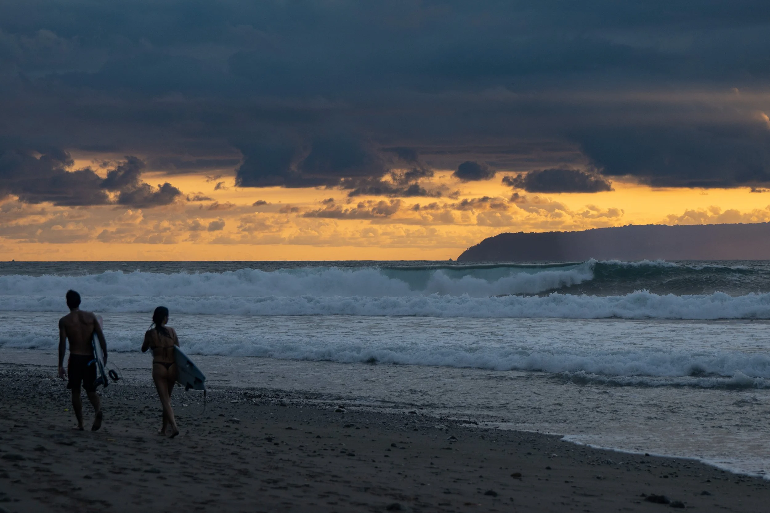 Two surfers walking on the beach with surfboards, preparing to surf at sunset, with dark clouds and a distant landmass in the background.
