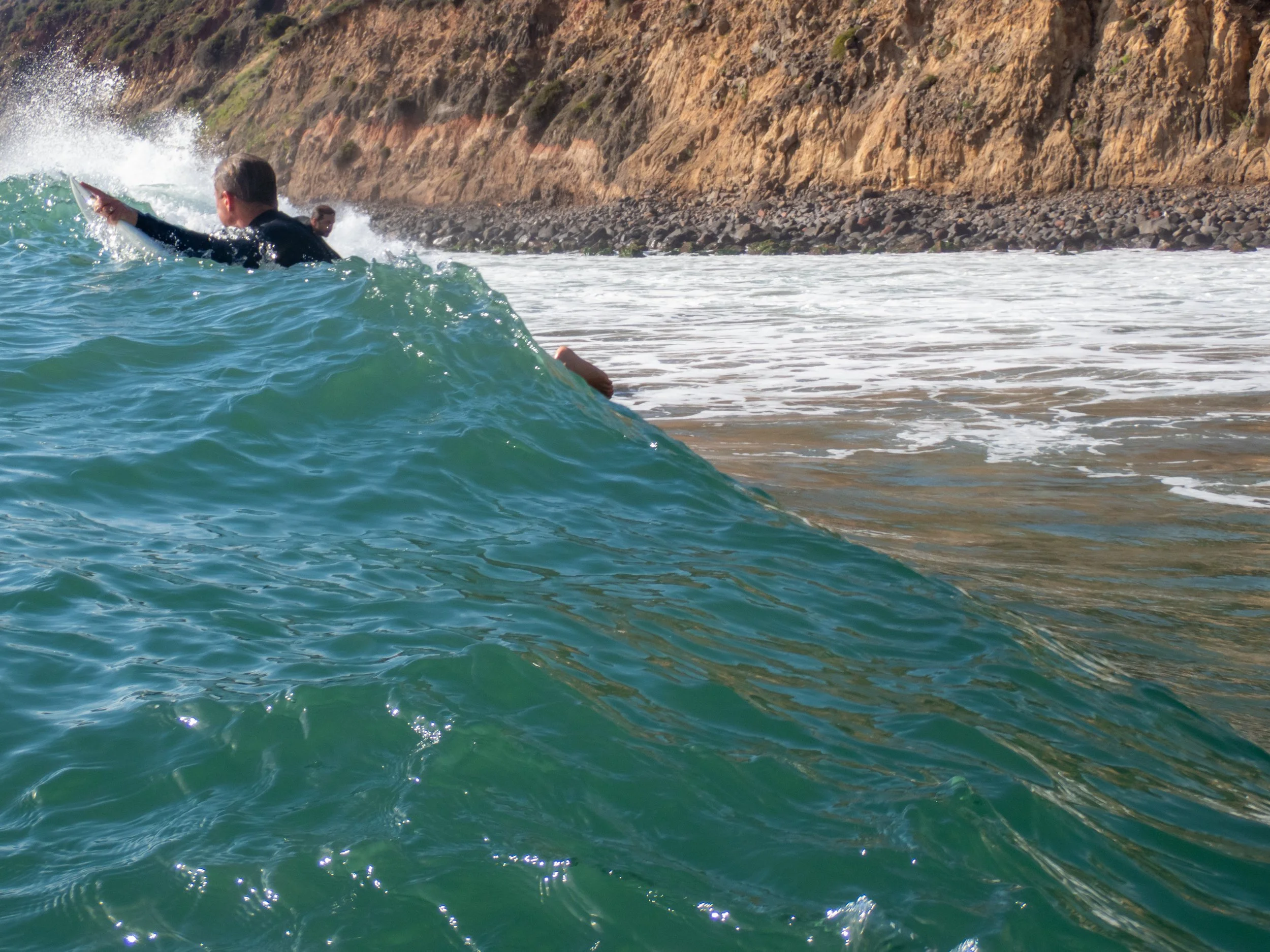 A person surfing on a small wave near a rocky coastline, with the ocean and cliffs in the background.