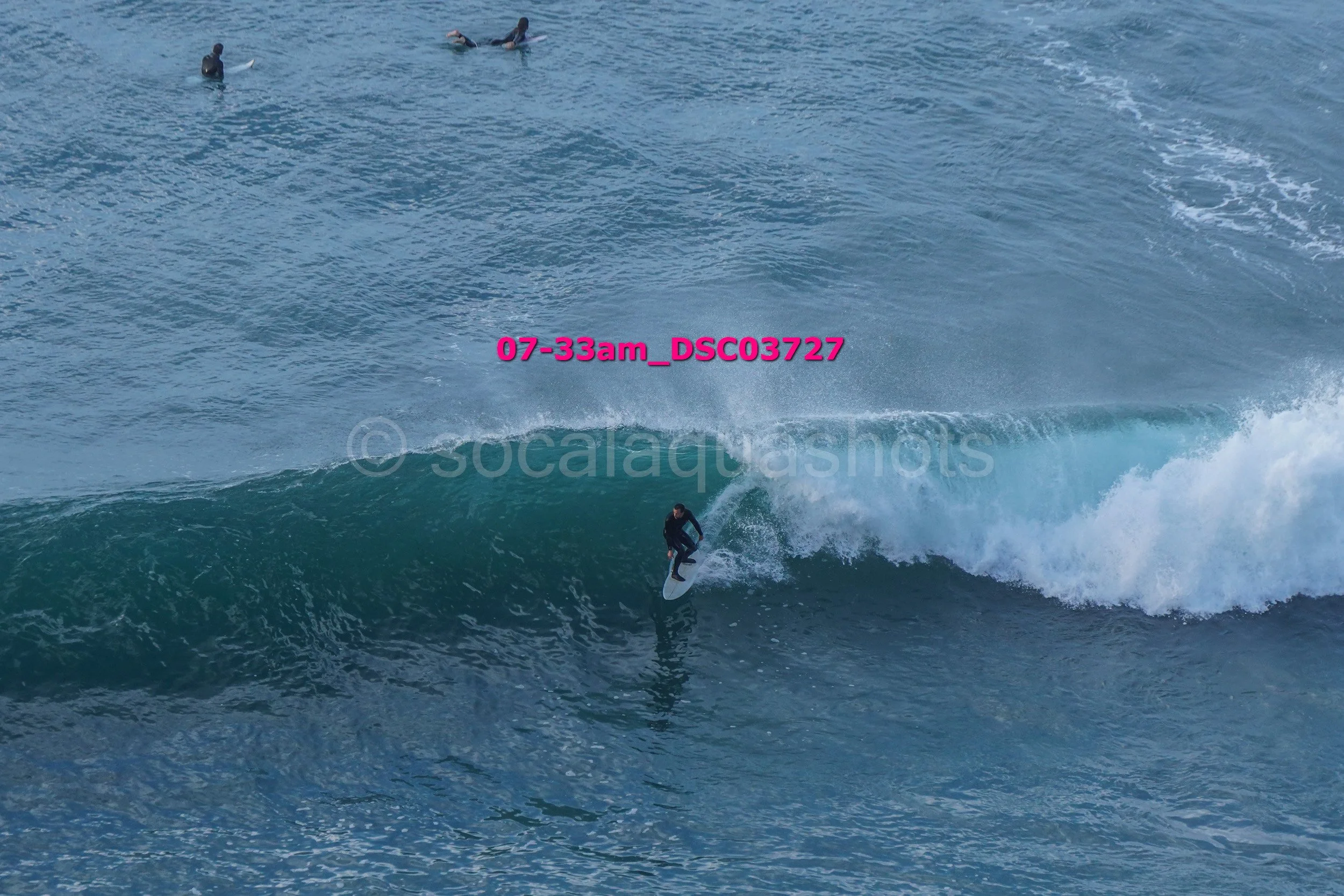 A person surfing on a large wave in the ocean, with three other people floating in the water nearby.