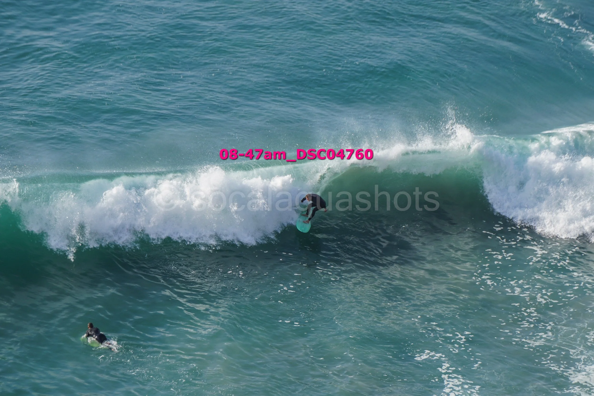 A surfer riding a large wave in the ocean, with another surfer paddling nearby.