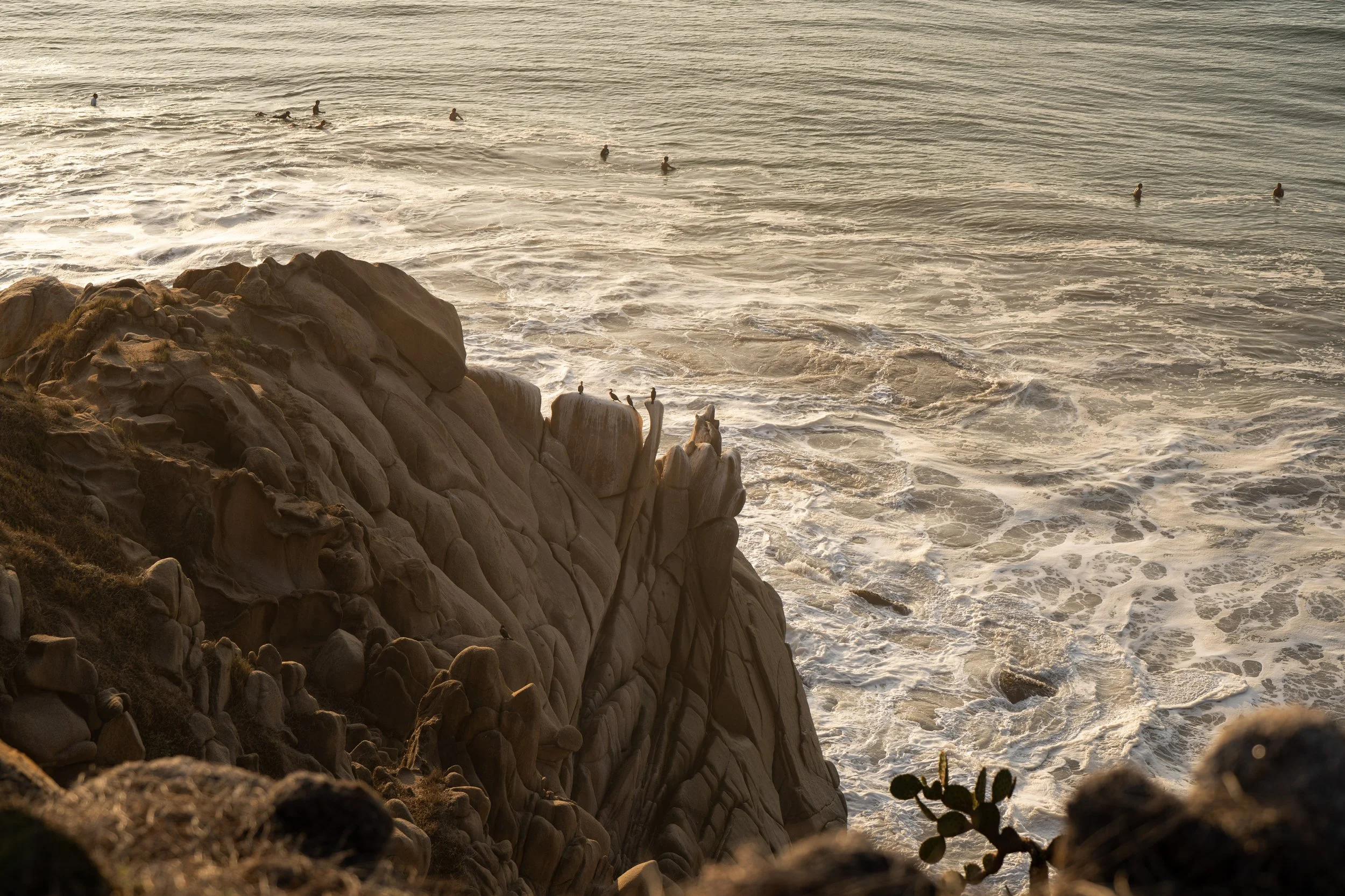Cliffs overlooking the ocean with people swimming in the water during sunset.