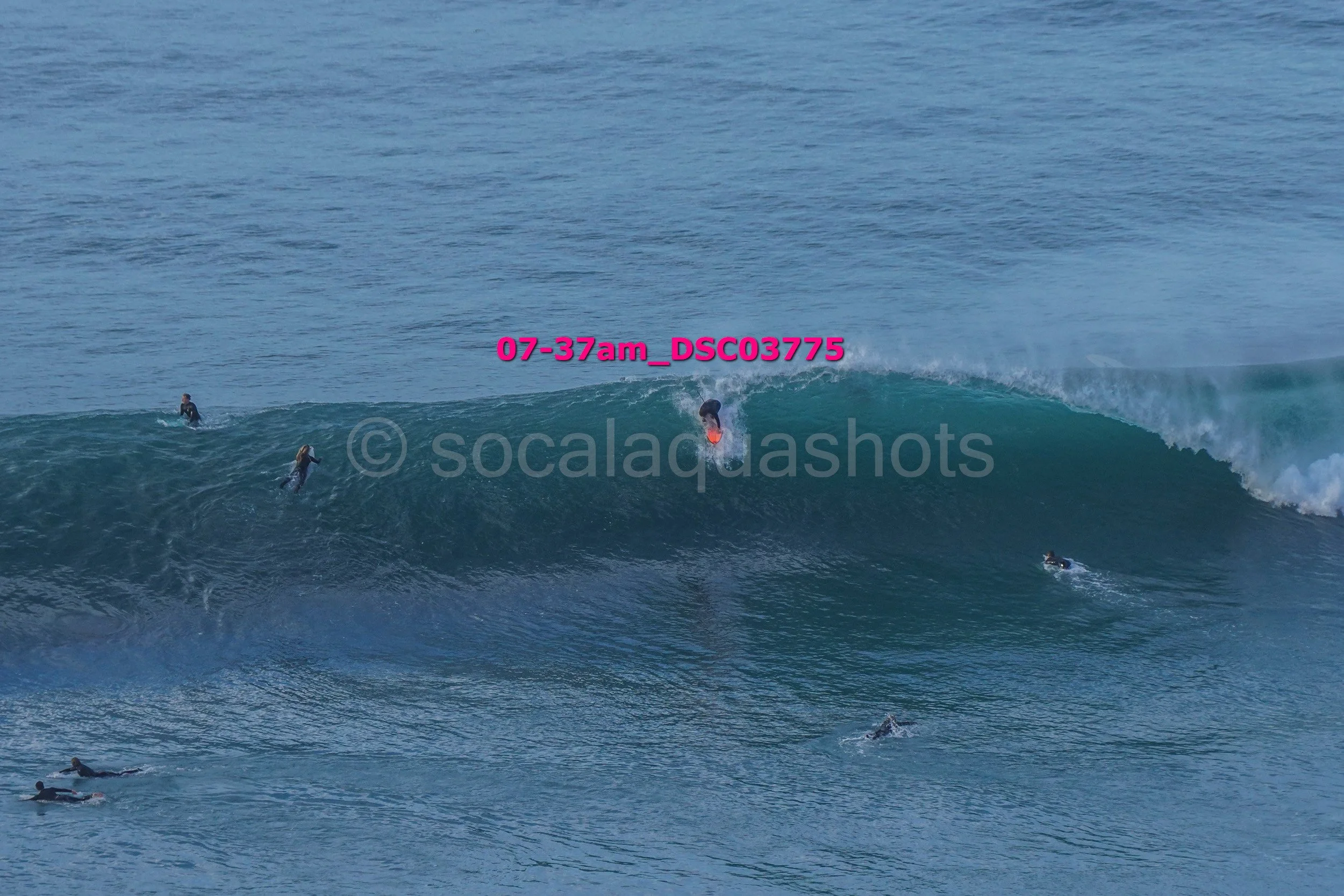 Surfers in the ocean, some riding a large wave, others paddling or waiting in the water.