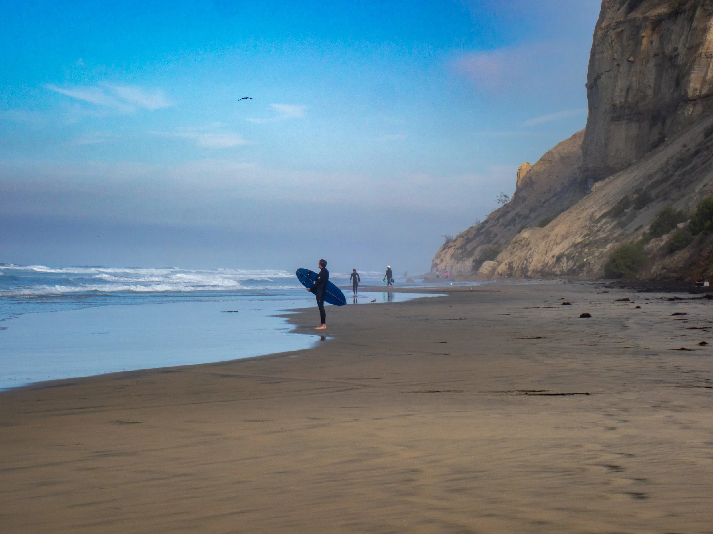 A beach with several surfers walking along the shoreline, some carrying surfboards, with a large cliff on the right and a blue sky overhead.