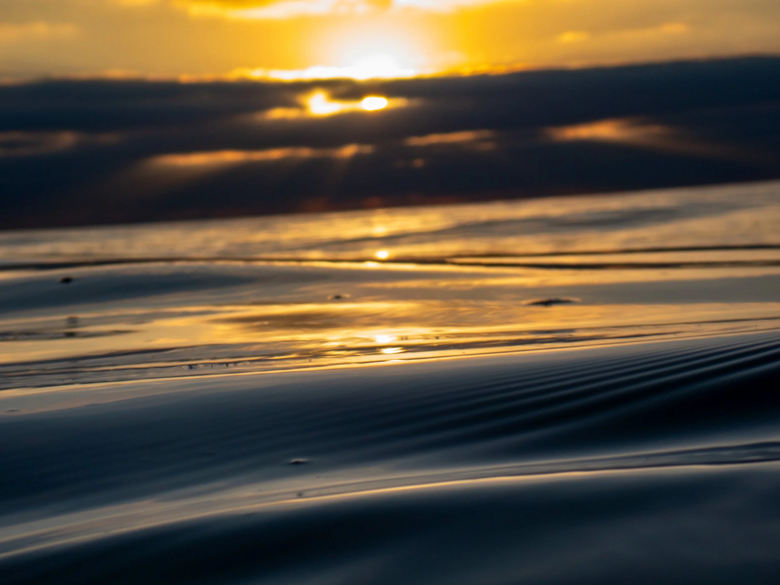 Close-up of water surface with gentle ripples, reflecting a sunset with a bright sun partially obscured by clouds.