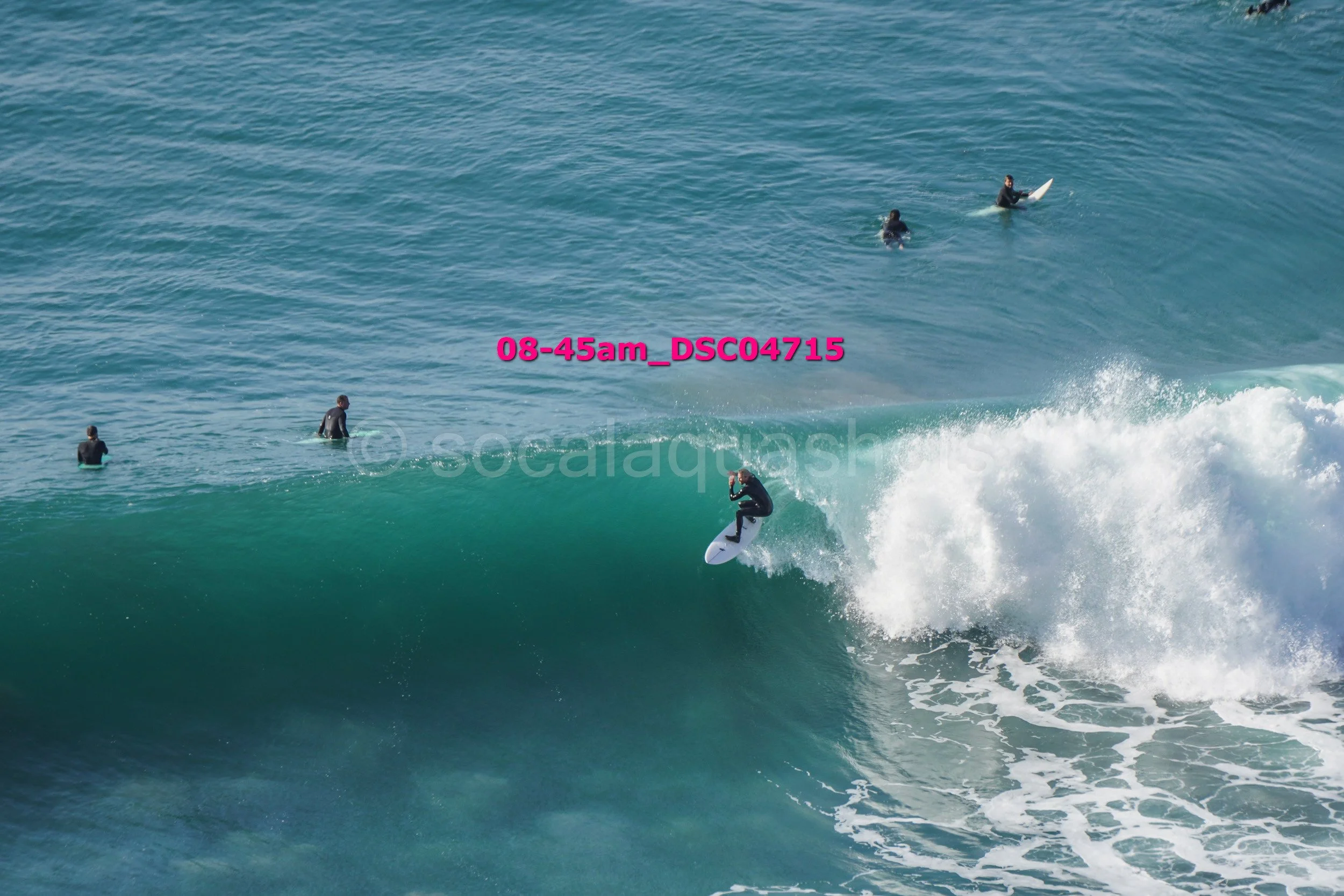 A person surfing on a wave in the ocean with four other people in the water, some on surfboards and some swimming.
