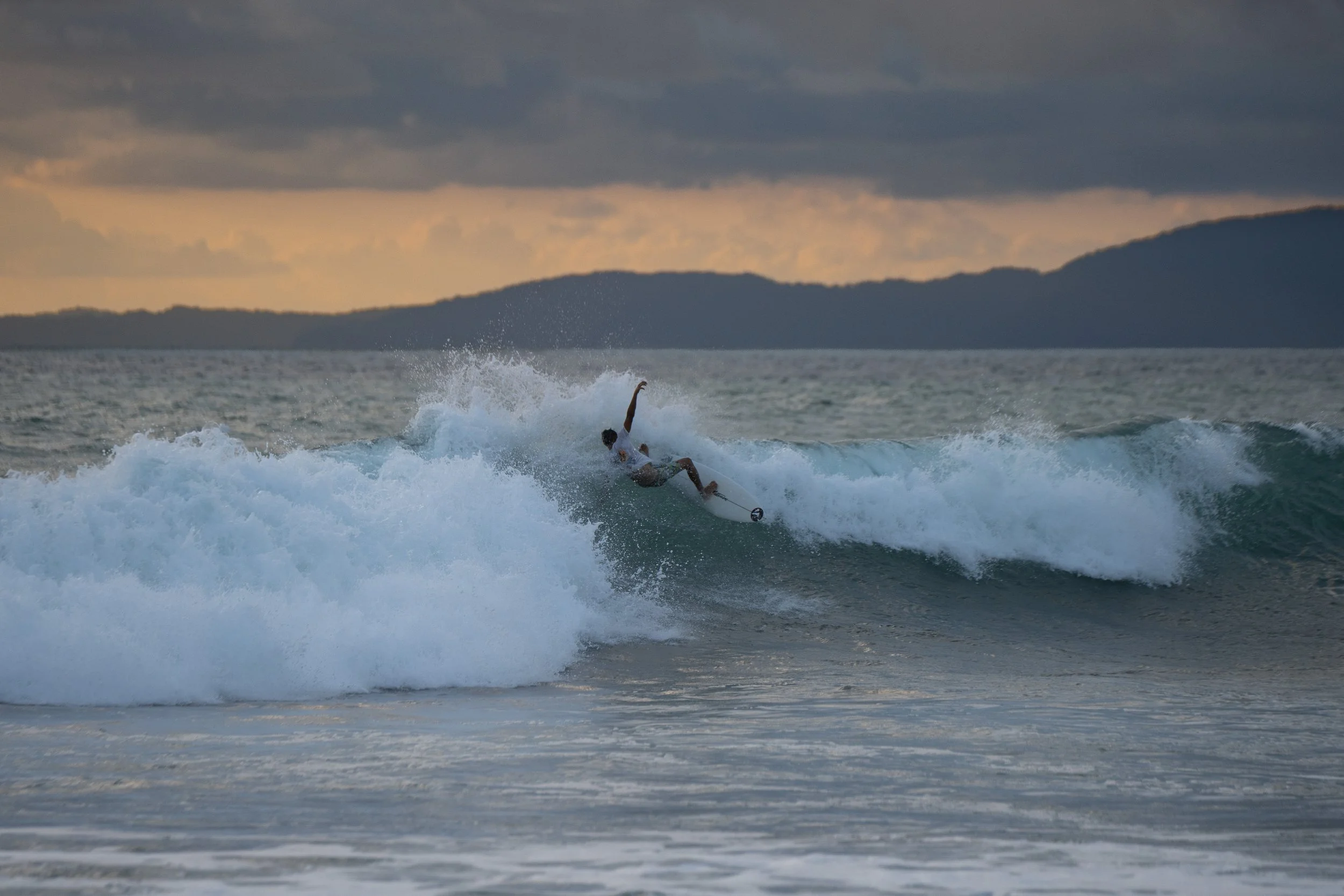 A person surfing on a wave in the ocean with a scenic mountainous landscape and cloudy sky in the background.