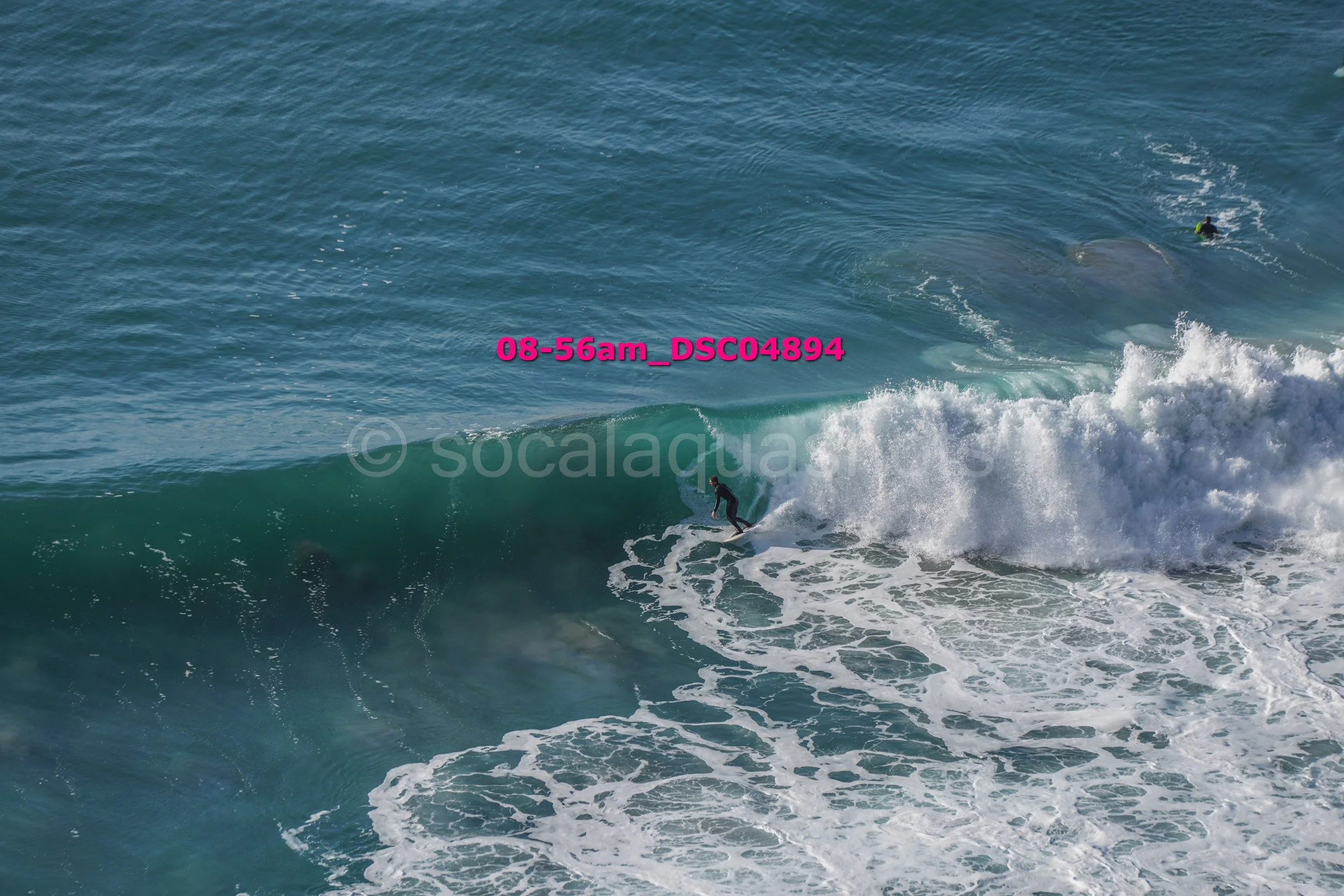 Surfer riding a wave in the ocean with another surfer visible in the background.