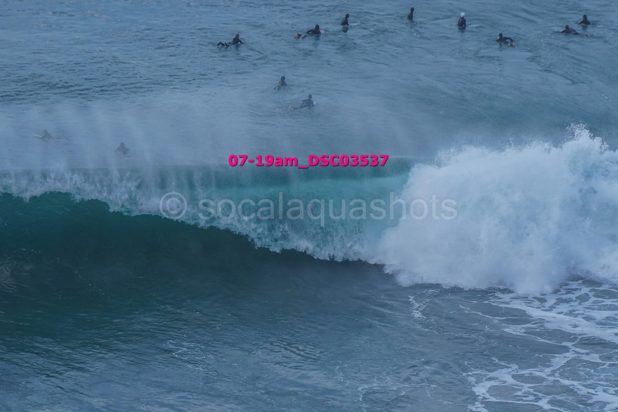 Multiple surfers in the ocean waiting for waves, with one wave in the foreground about to break.