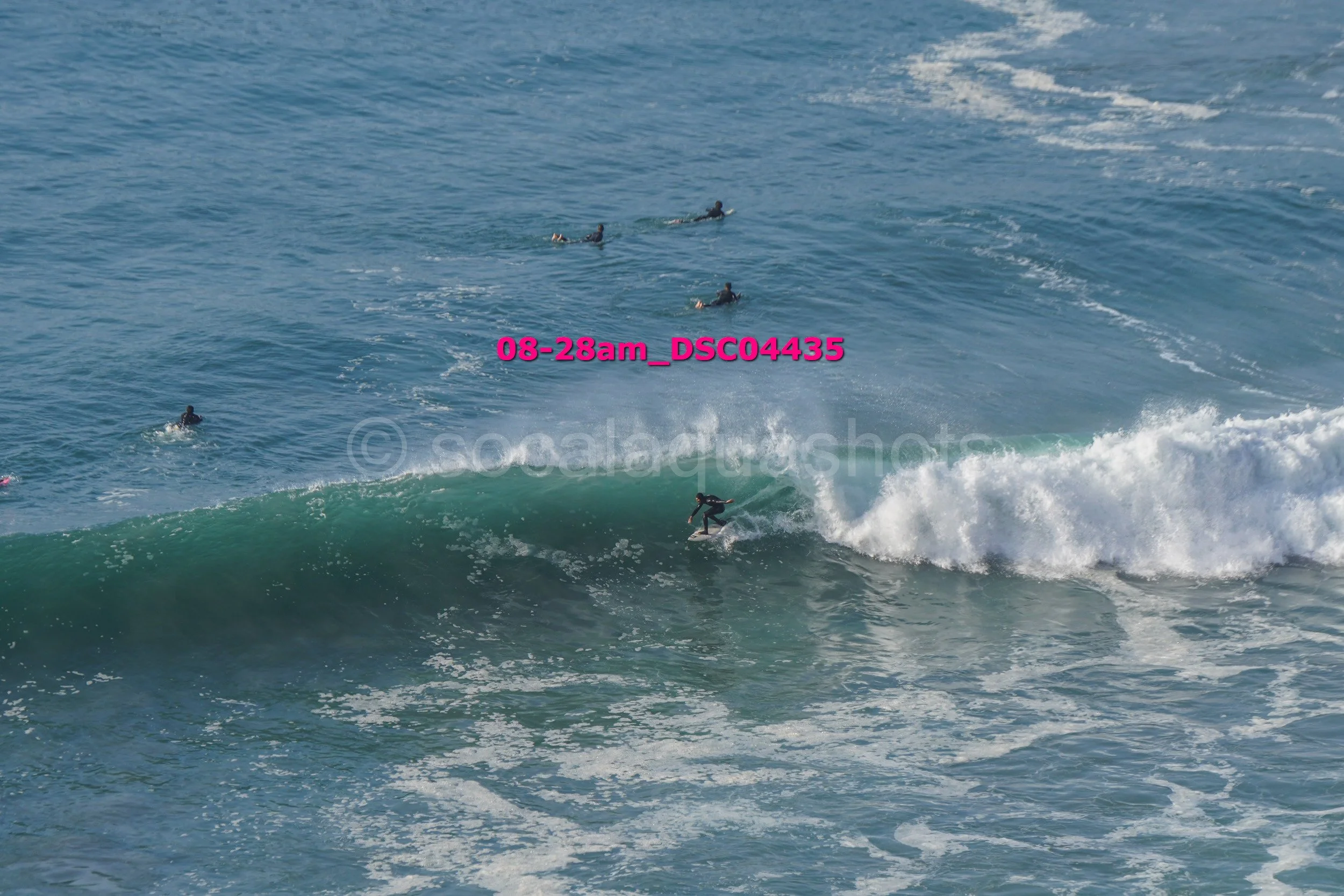 Surfer riding a wave in the ocean with several people swimming or surfing in the background.