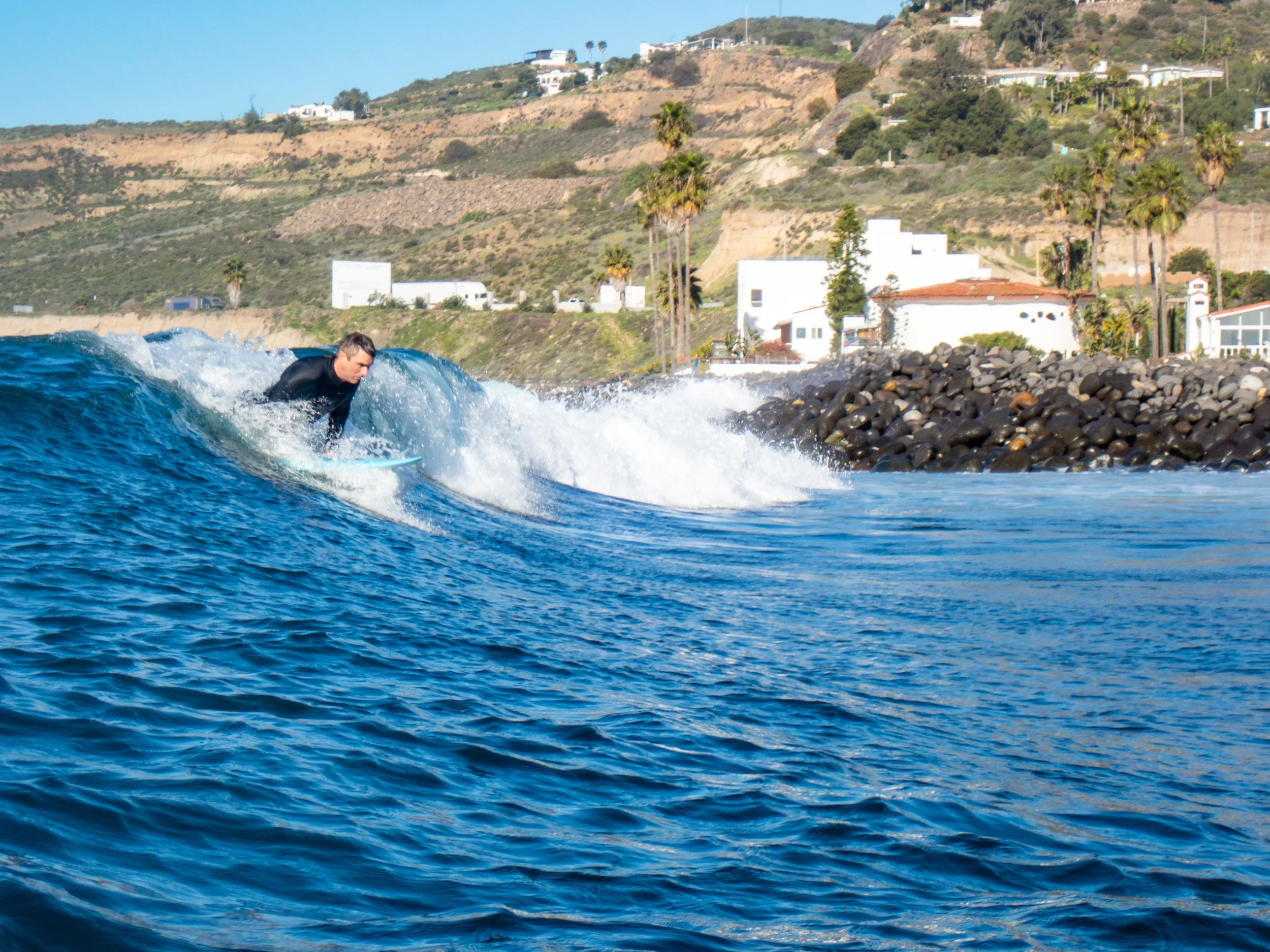 A person surfing on a wave with a coastal hillside and houses in the background.