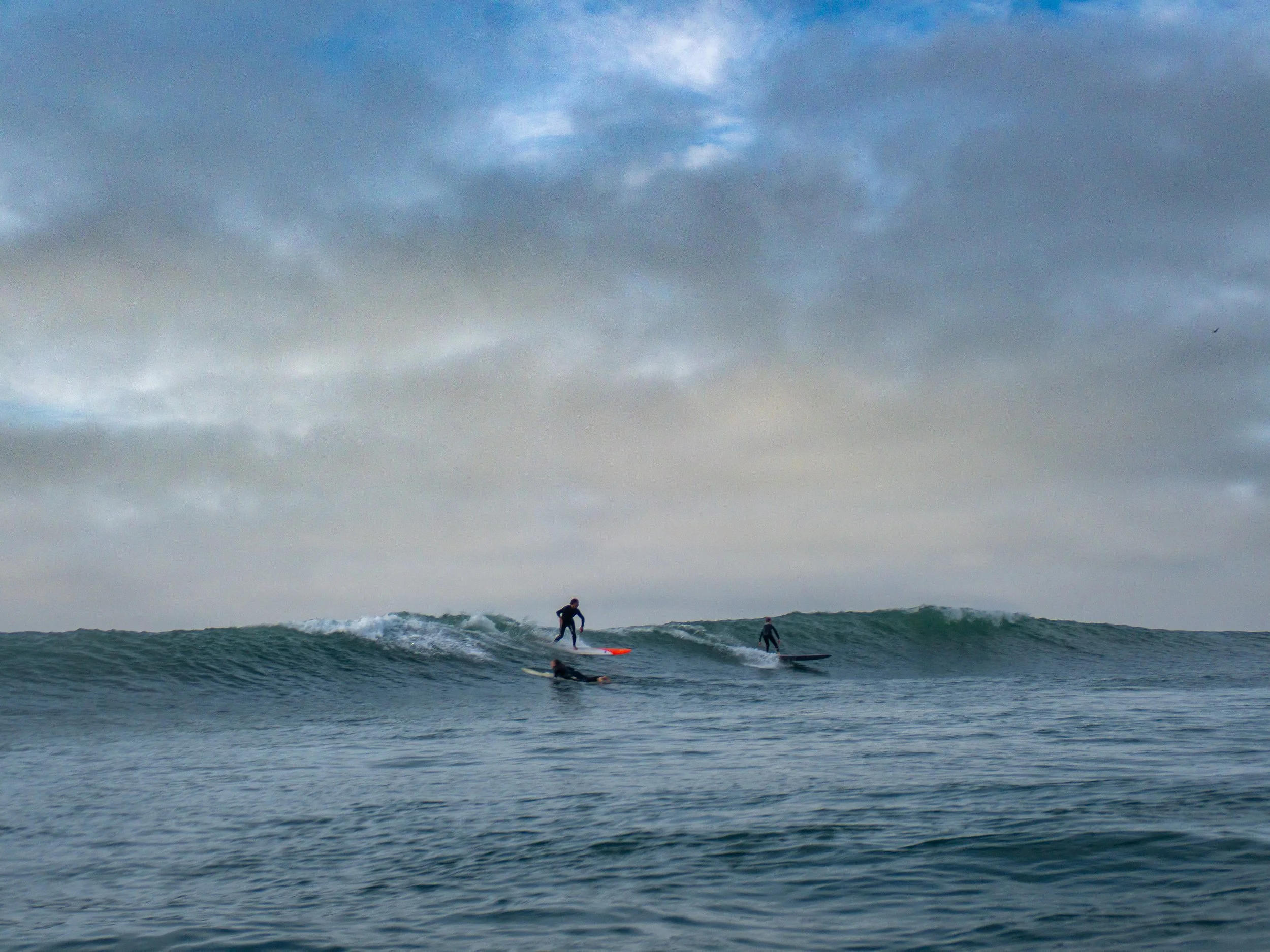 Three surfers riding and paddling on ocean waves under a cloudy sky.